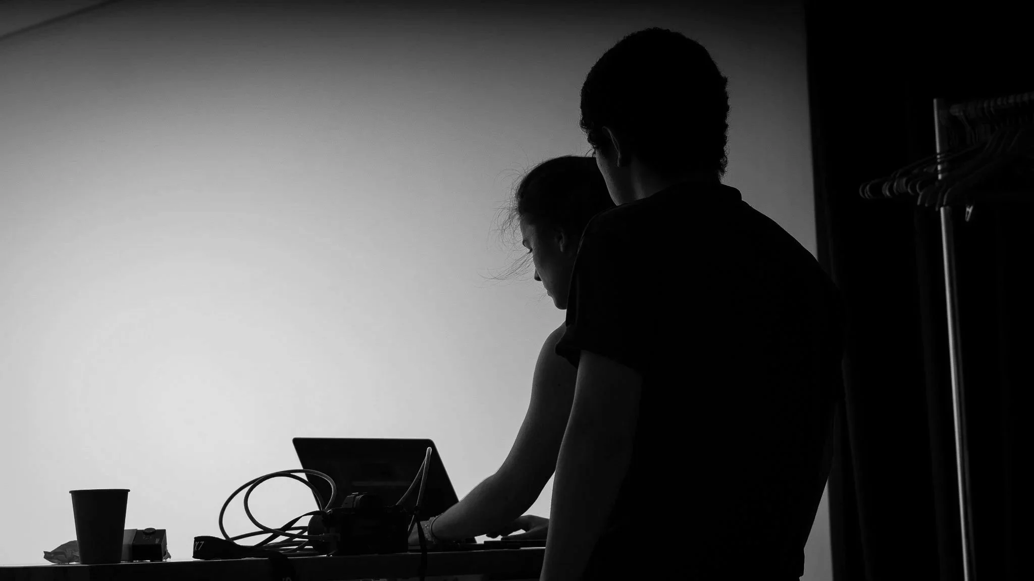 Silhouettes of a man and woman working together at a table with electronic equipment and a laptop in a room with a coat rack.