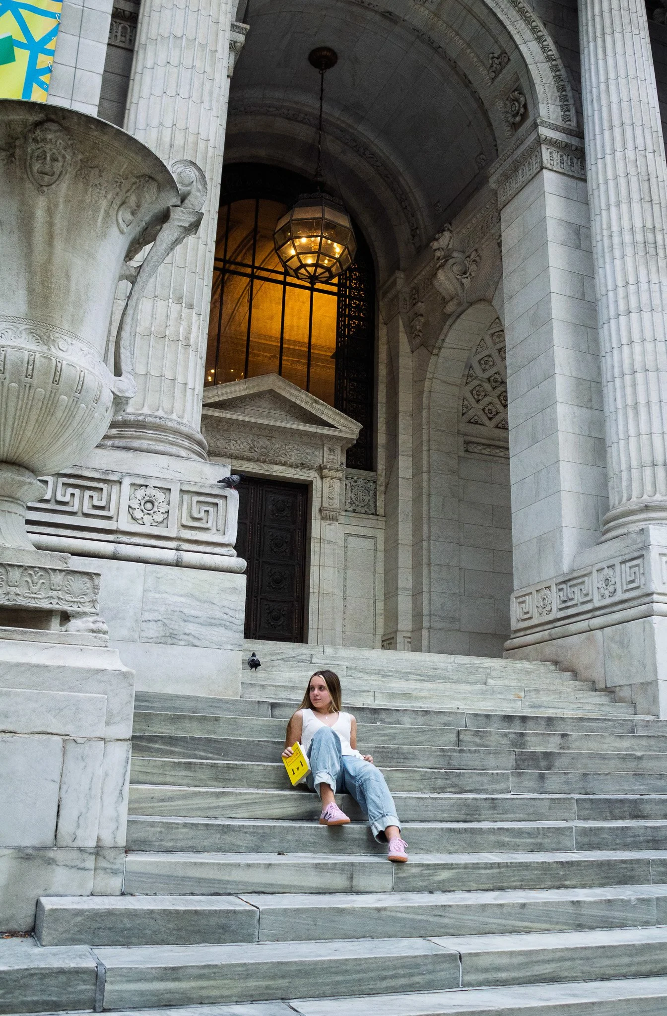 A young woman sitting on the marble steps of a large, classical-style building with tall columns and ornate architecture, holding a yellow booklet and a snack, with pigeons nearby.