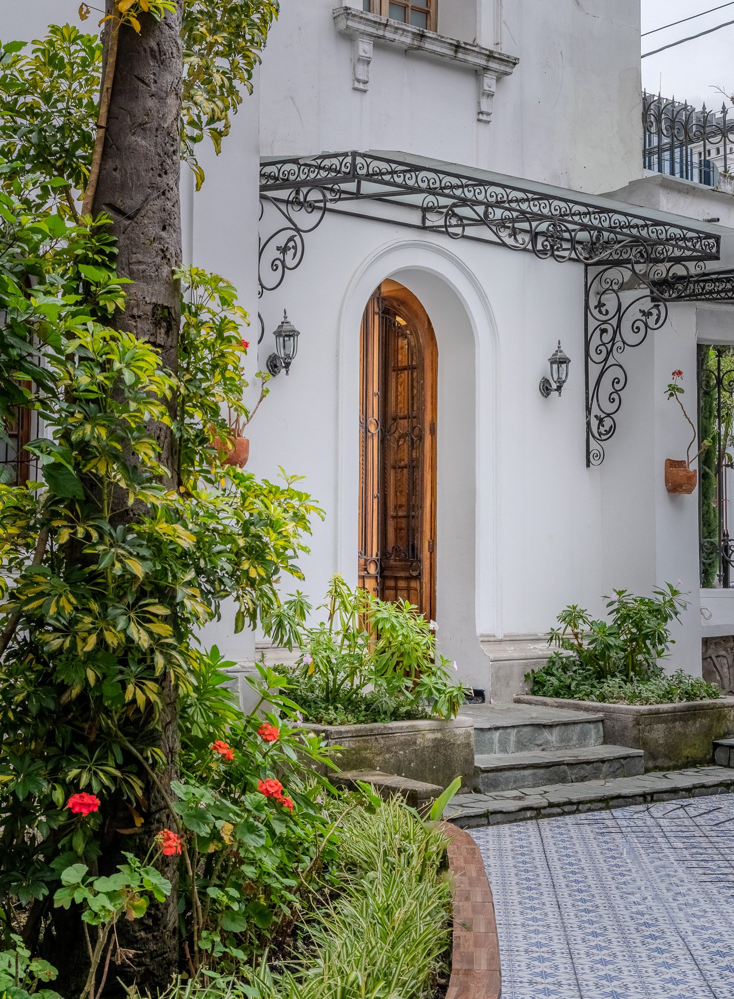 The image shows the entrance to a white house with wooden door and arched doorway, surrounded by green plants and flowers, including pink and red blooms. A tree with textured bark is visible on the left side, and stone steps lead up to the door. Deco