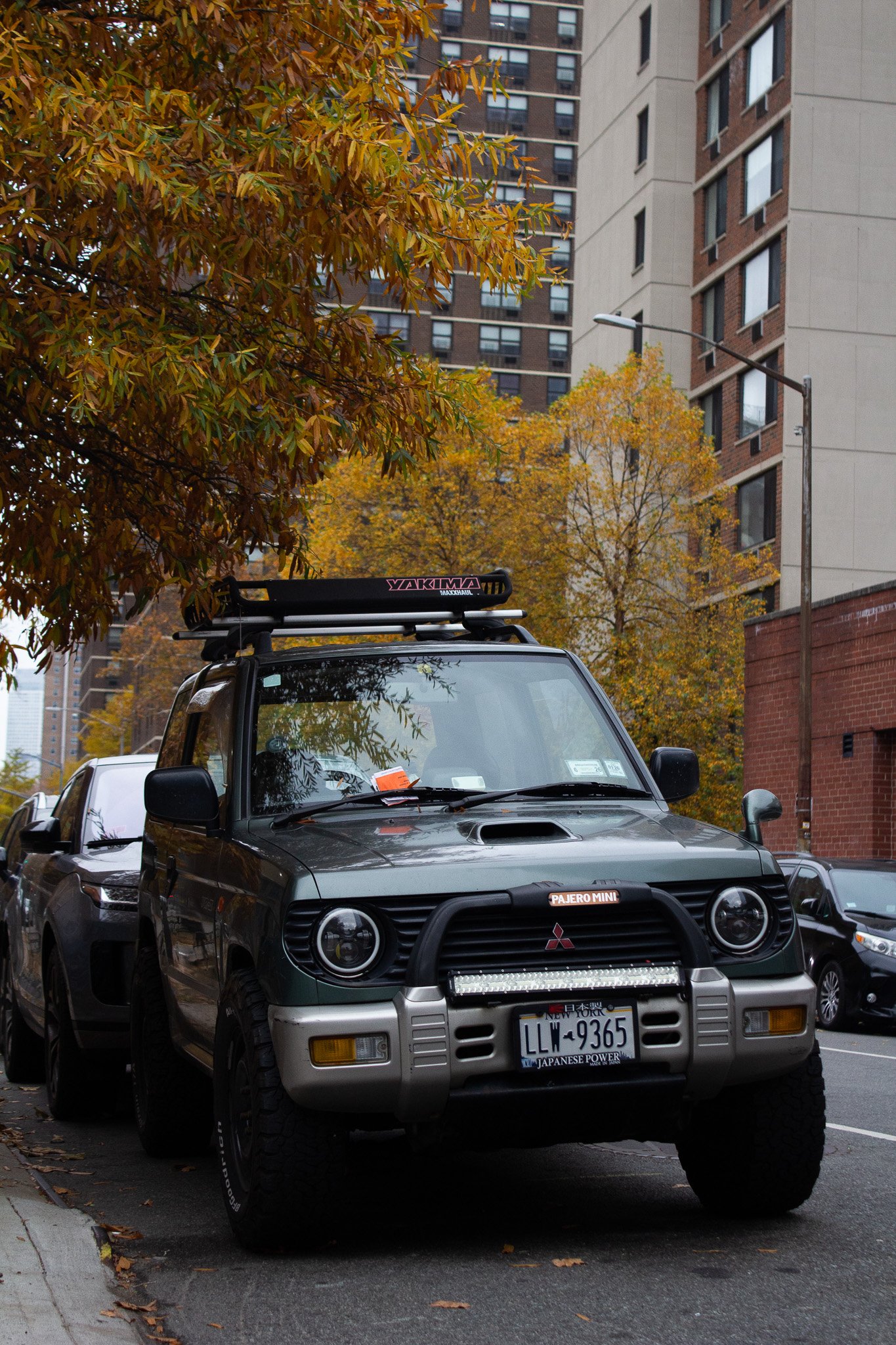 A black Mitsubishi Pajero Mini SUV parked on a city street with trees and tall buildings in the background. The vehicle has a roof rack and a front bumper guard, with a New York license plate and various stickers.
