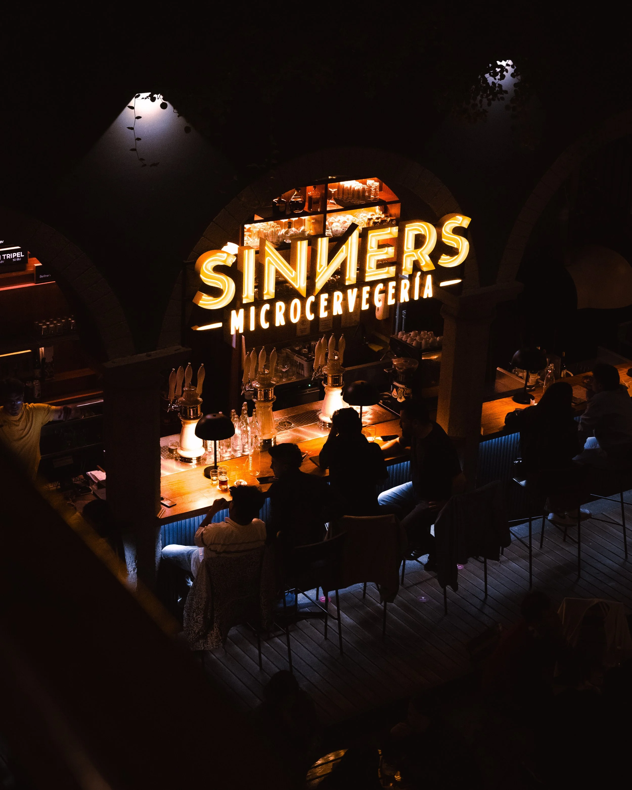 Nighttime scene of a bar or restaurant named SINNERS MICROCERVERCIA with illuminated sign, bar counter with illuminated taps, glasses, and bottles, and customers sitting and socializing inside, under a dark decorative ceiling with hanging plants.