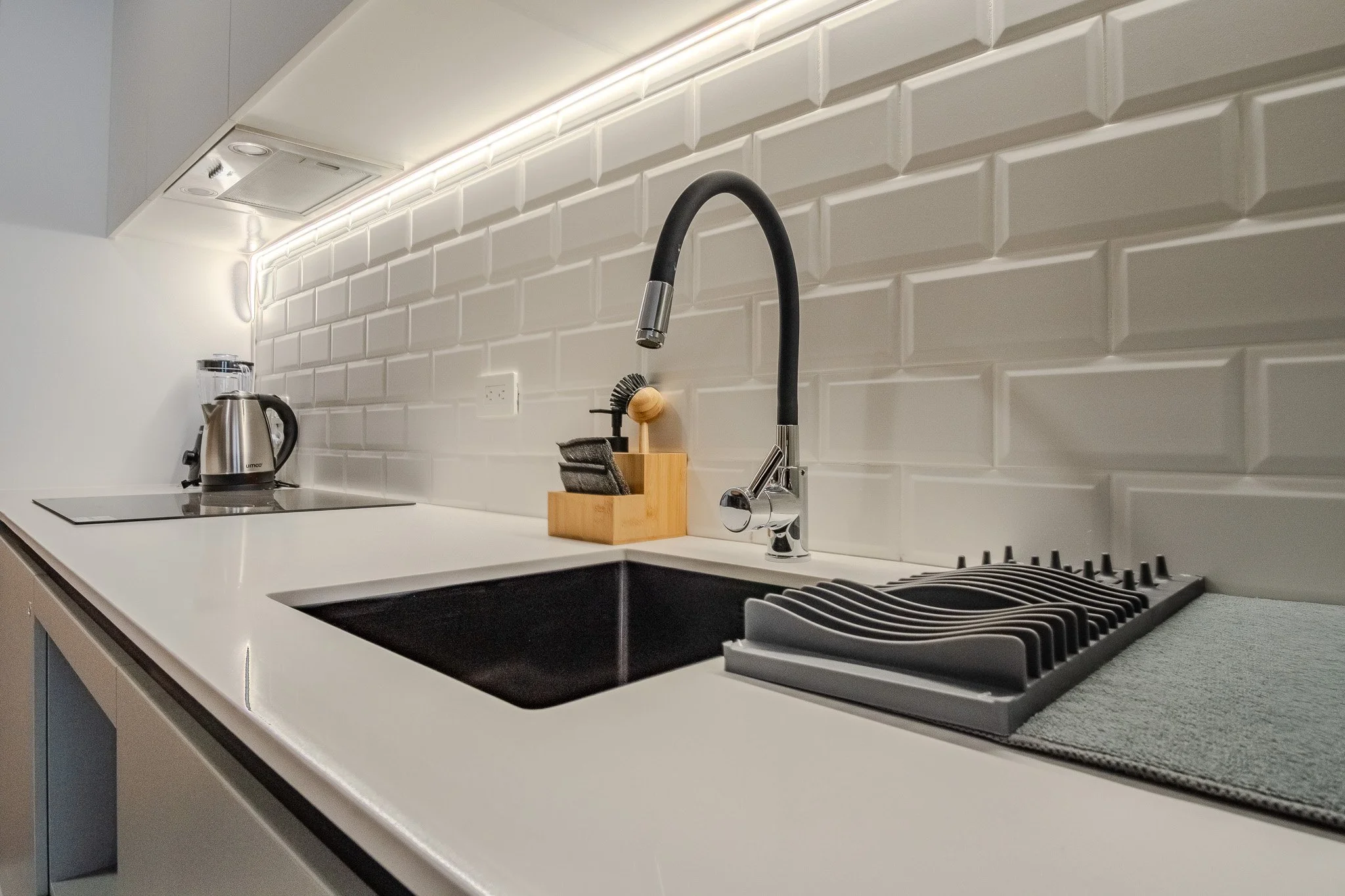 Modern kitchen counter with a black sink, a black and silver faucet, a drying rack, and a tea kettle, with white cabinetry and a white tiled backsplash.