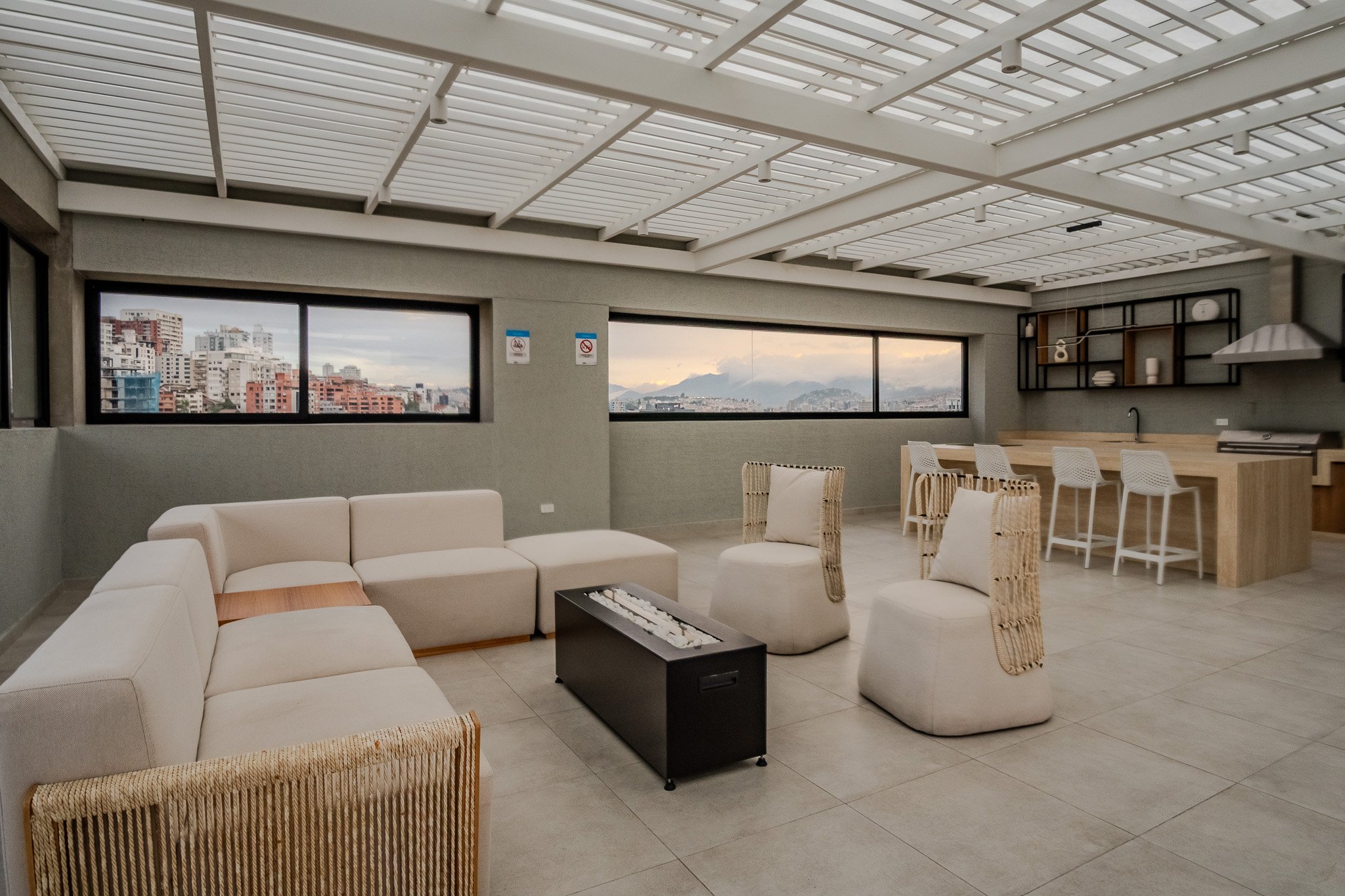 Modern indoor living space with beige sectional sofa, white armchairs, wooden coffee table with a black decorative piece, and large windows showing cityscape and mountains in the distance.