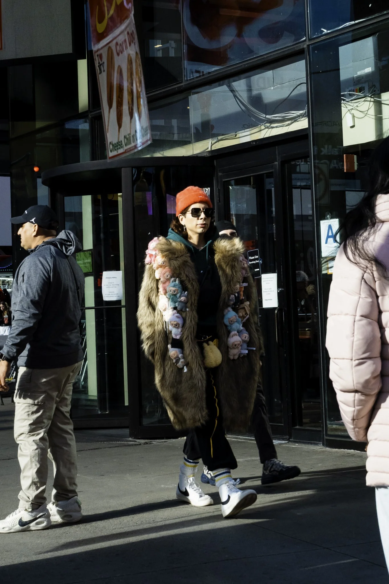 A woman in a fur coat decorated with plush toys, wearing sunglasses and a red beanie, walking outside an entrance with glass doors, with other people around and a McDonald's sign overhead.