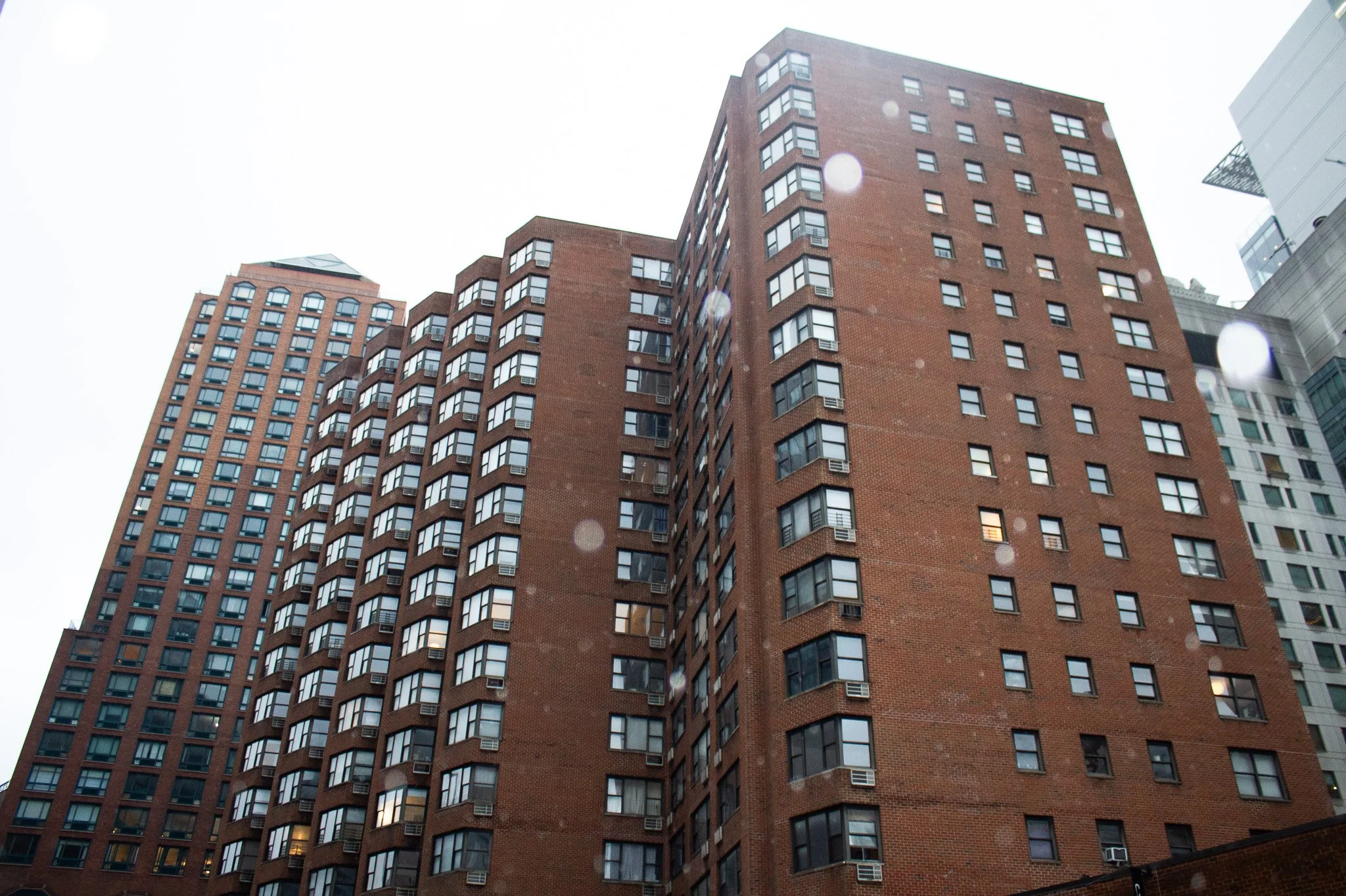 Tall brick apartment buildings in an urban area with numerous windows, some with air conditioning units, under an overcast sky.