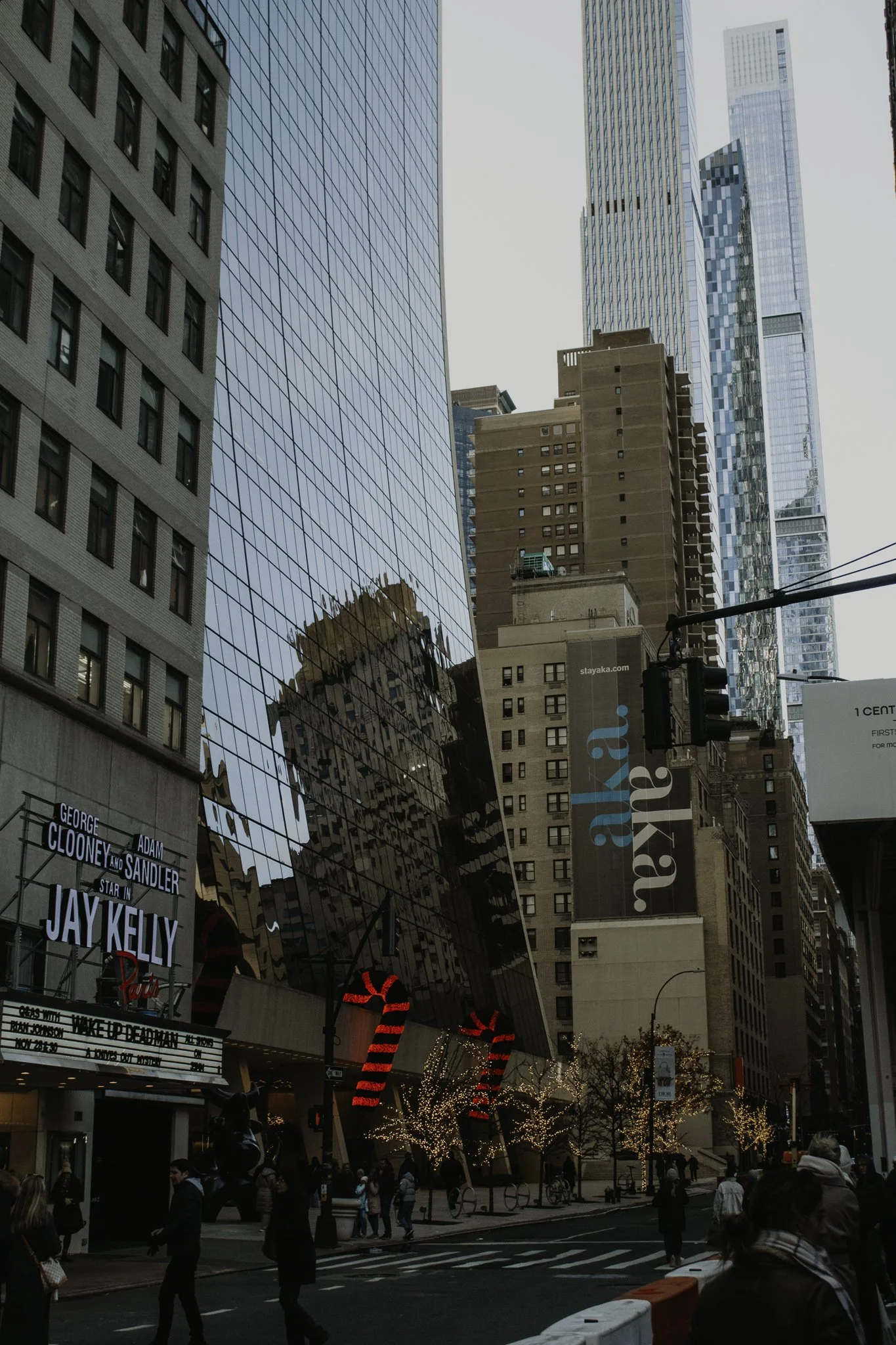 A busy city street scene with tall glass and concrete buildings, some with holiday decorations and lights, and people walking on the sidewalk.