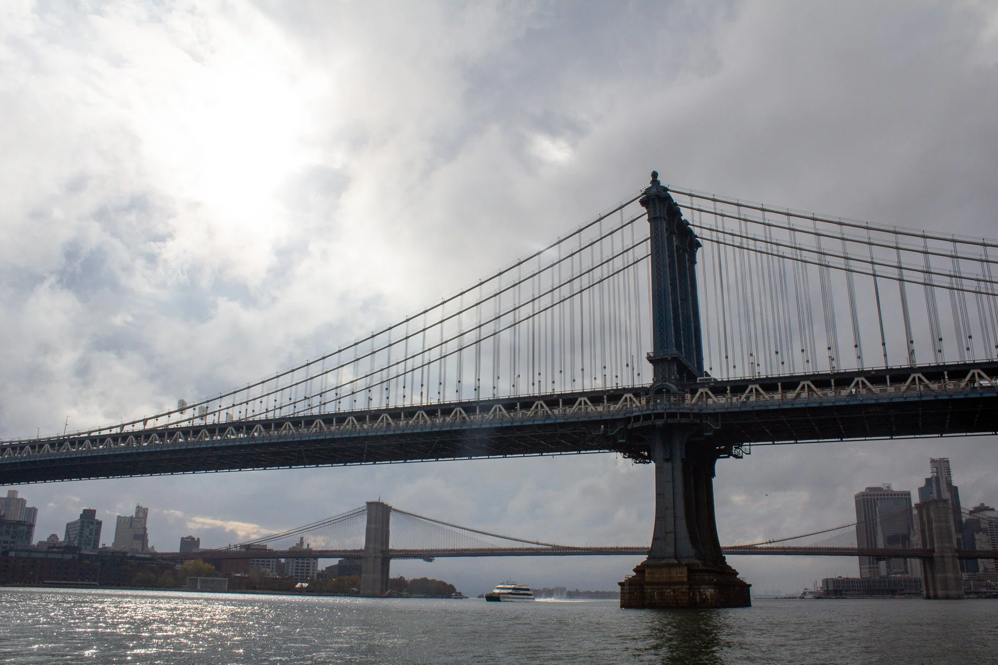 View of the Brooklyn Bridge and Manhattan Bridge over the East River in New York City, with buildings and a boat in the water under a cloudy sky.