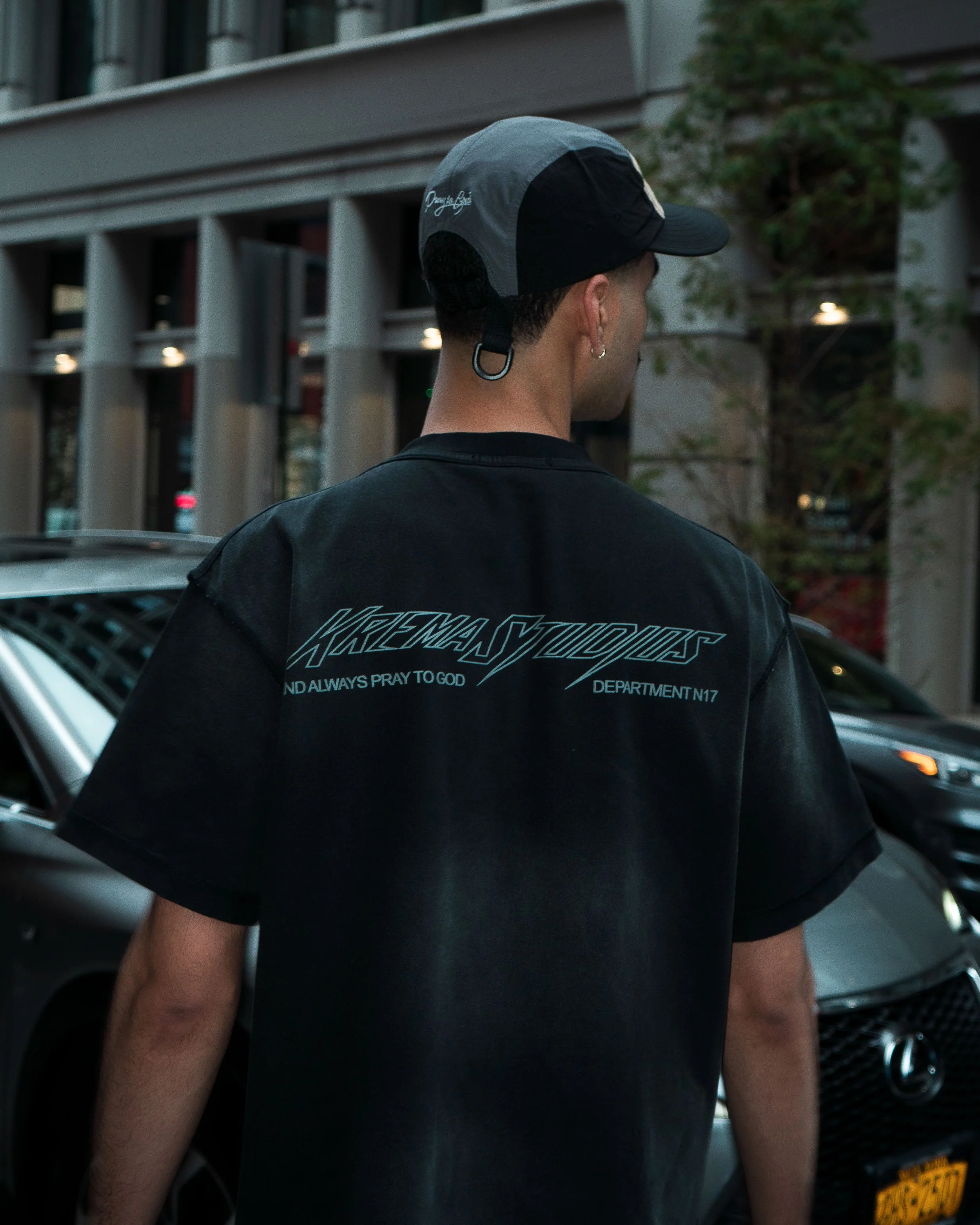A man wearing a black T-shirt and a gray and black cap stands outdoors in front of a row of parked cars, with a building in the background. The T-shirt has stylized text on the back that says "KREMA vystuz" and additional smaller text.