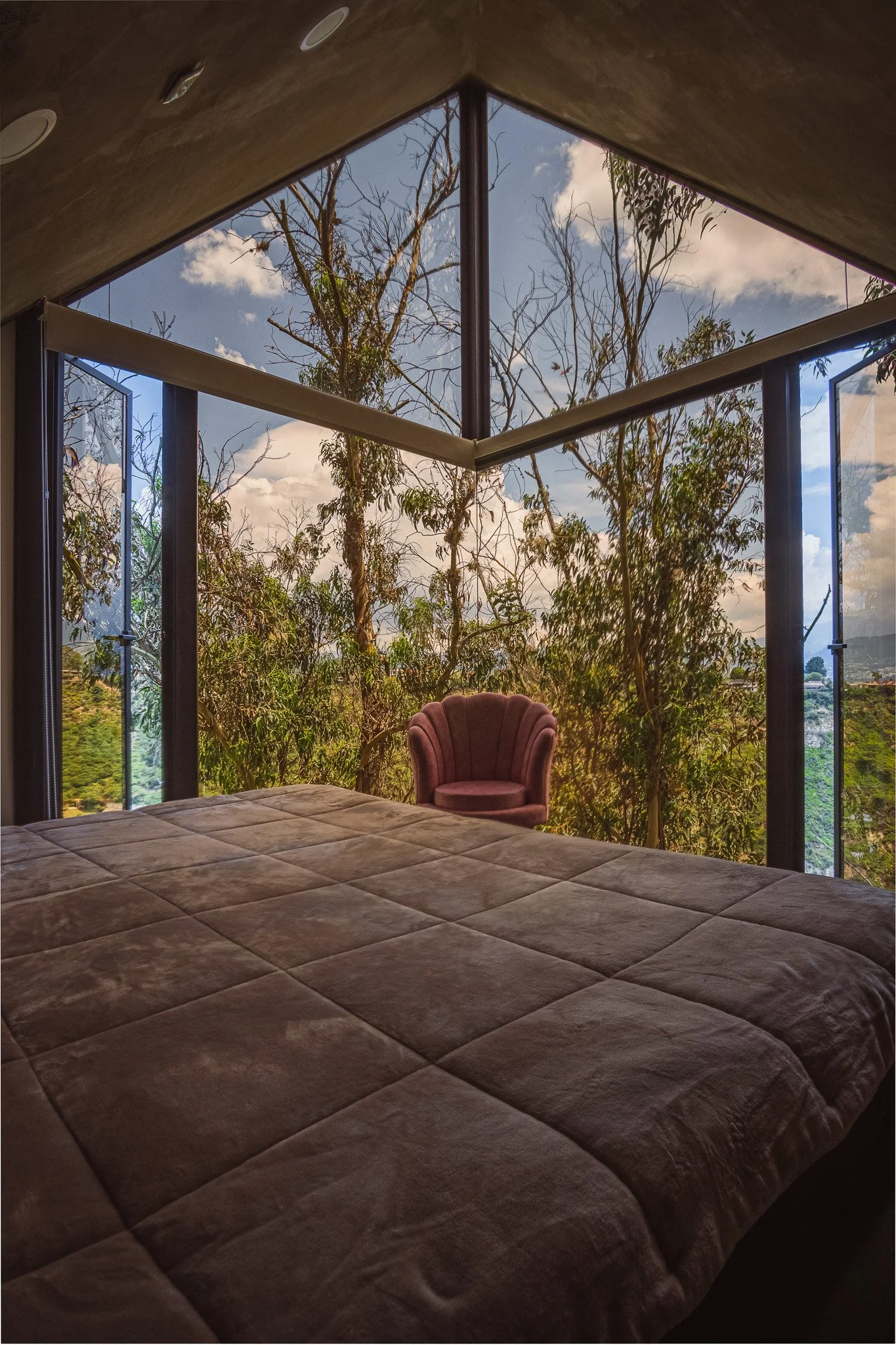 A bedroom with a bed in the foreground, a glass wall showing trees and a cloudy sky outside, and a pink armchair near the window.
