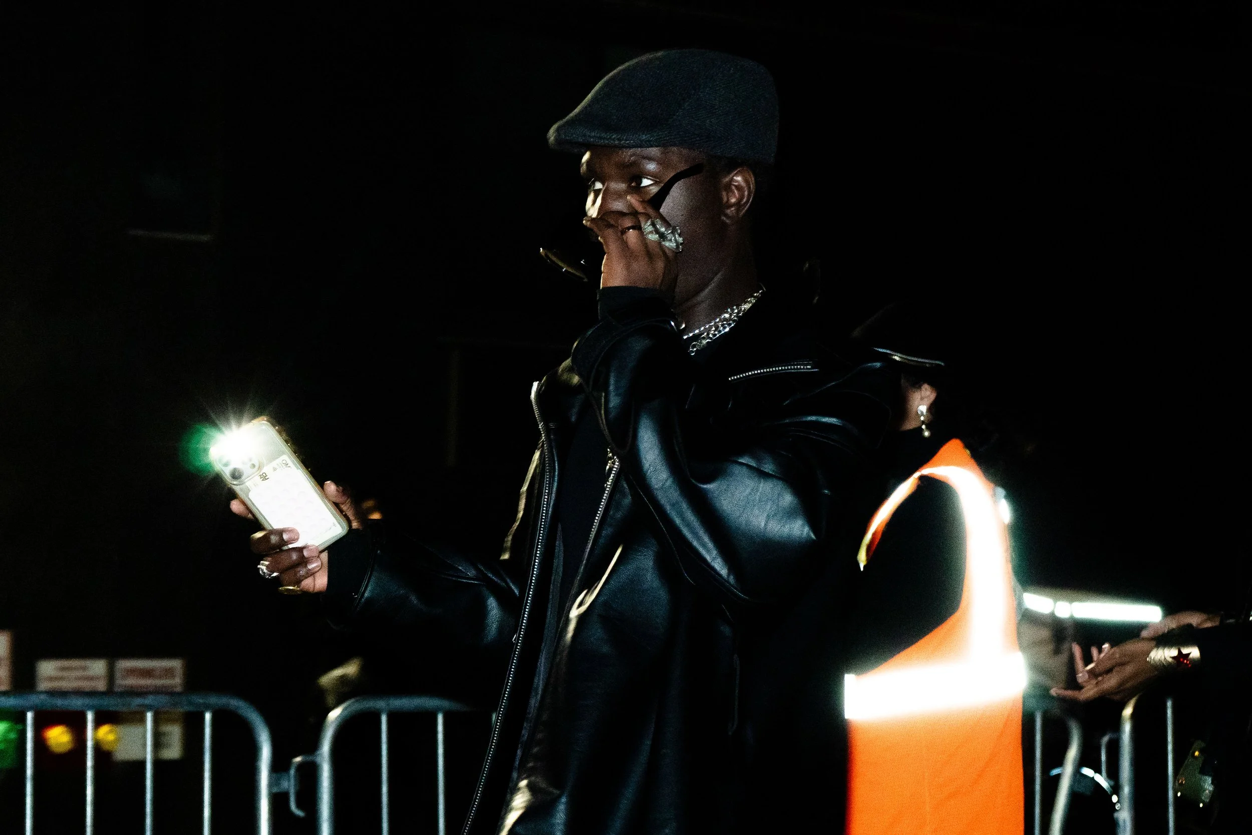 A person wearing a gray flat cap, glasses, multiple rings, silver jewelry, and a black leather jacket is talking on a phone and holding a smartphone with the camera flash on, at night, with a woman in an orange safety vest standing nearby, behind a m