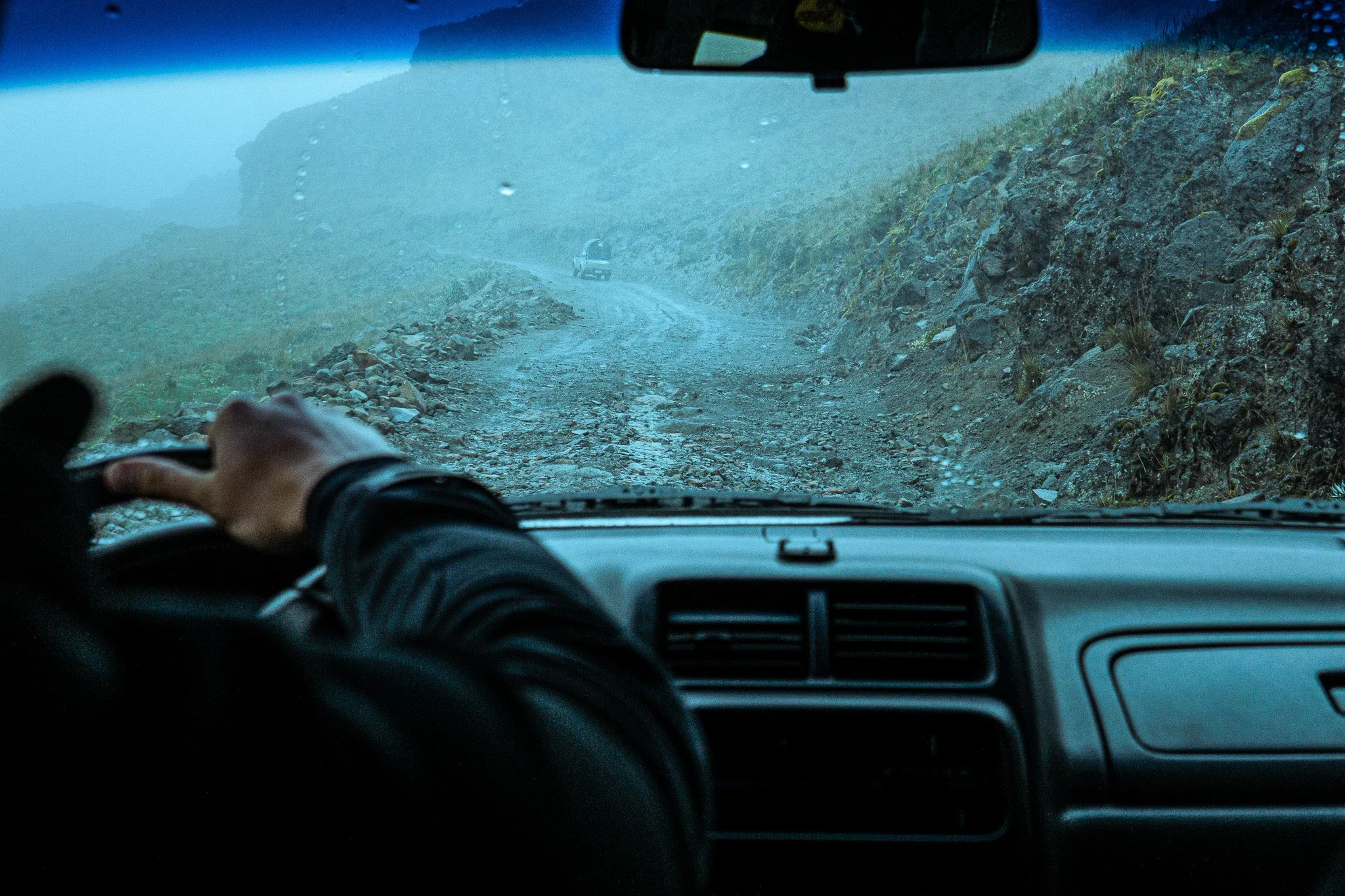 Interior view from inside a vehicle showing a rugged, rocky dirt road ahead, surrounded by mountains and fog, with a driver’s hand on the steering wheel.