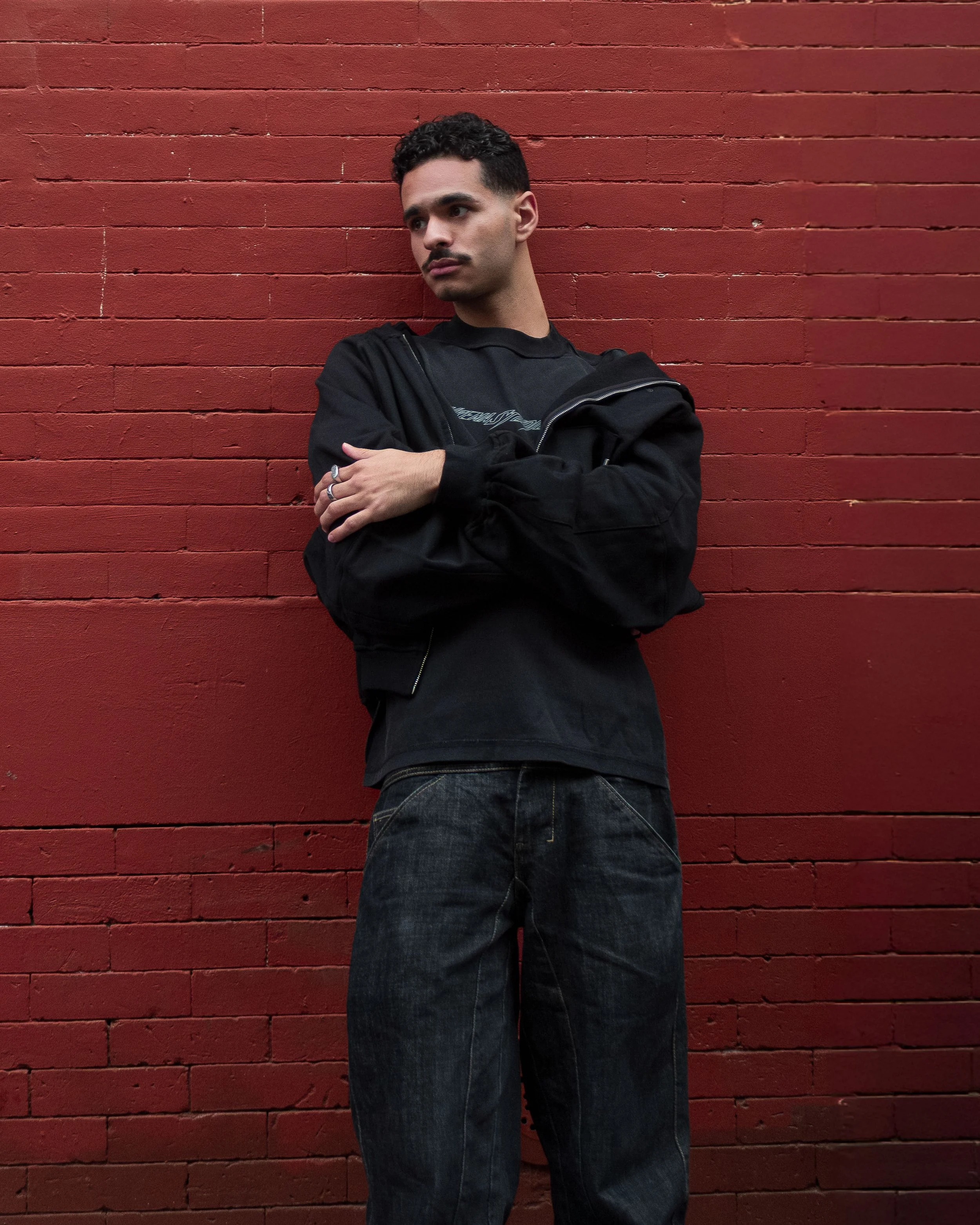 A young man with short dark hair and a mustache posing against a red brick wall, wearing a black jacket, black shirt, and dark jeans.