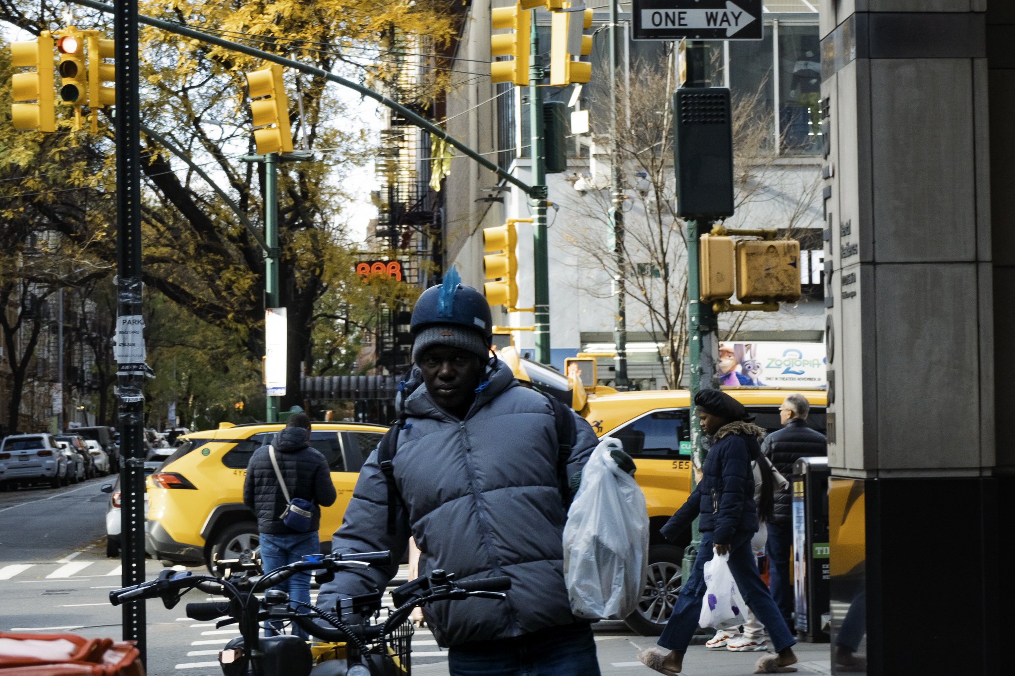 City street scene with people walking on the sidewalk, yellow taxis, and traffic lights. Some trees with autumn leaves are visible. There's a person in a dark jacket and beanie in the foreground, and others crossing or standing nearby. Buildings and 