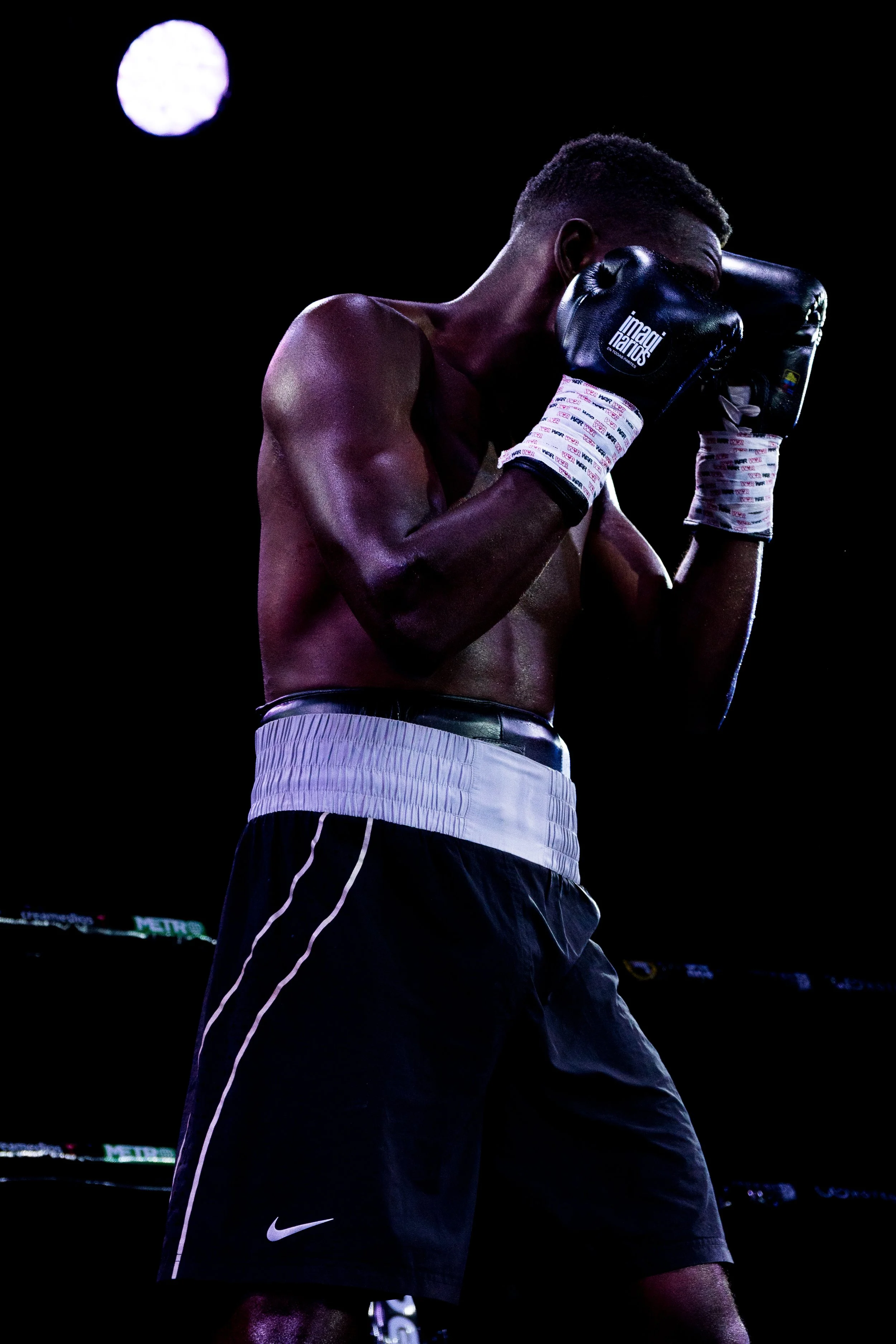 Boxer wearing black gloves in a fighting stance, shirtless, wearing black shorts with white stripes, standing in a dark arena with a bright moon in the background.