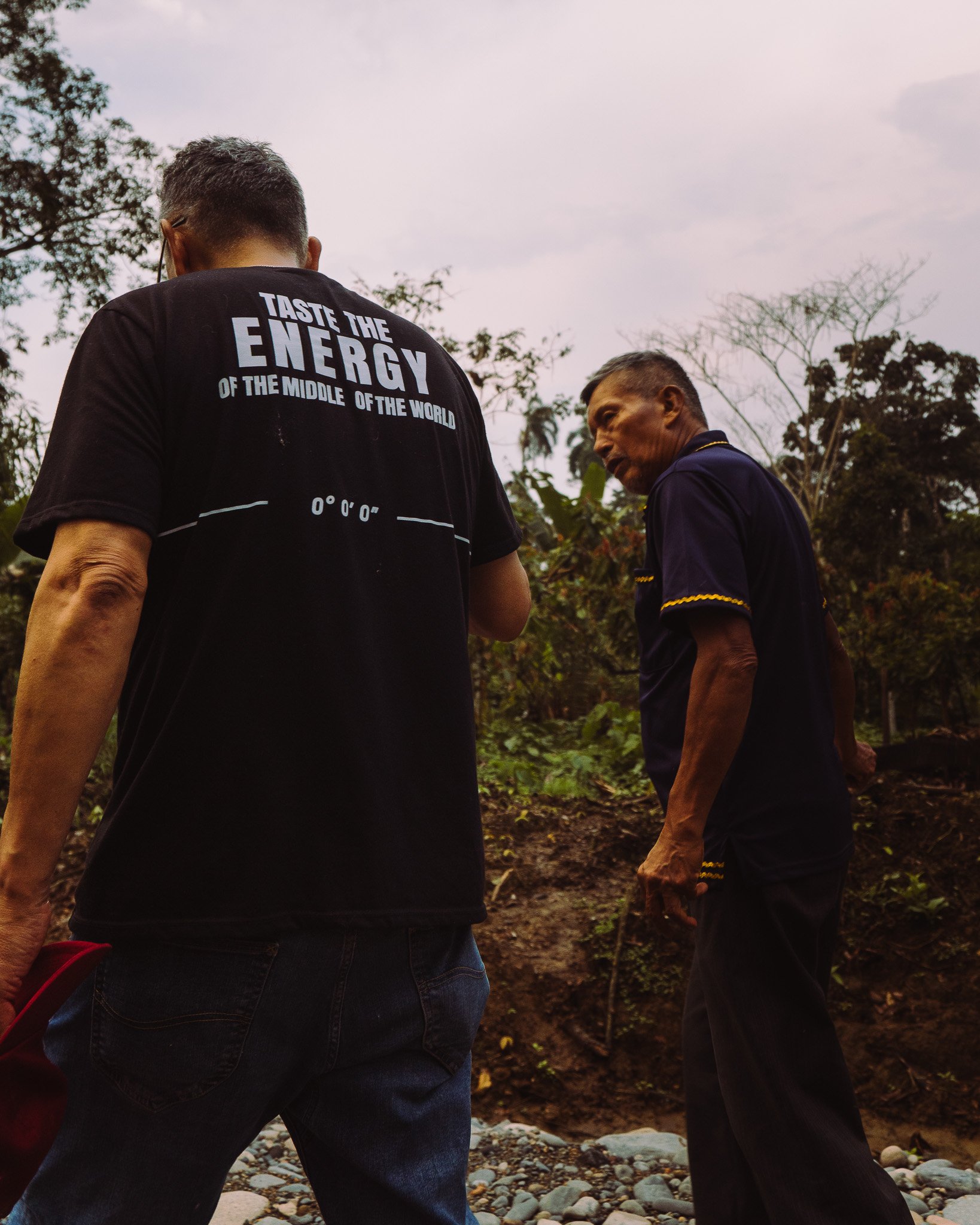 Two people walking outdoors on rocky terrain, one wearing a black T-shirt with white text that says 'Taste the energy of the middle of the world,' and the other in a dark shirt with yellow trim, surrounded by trees and an overcast sky.