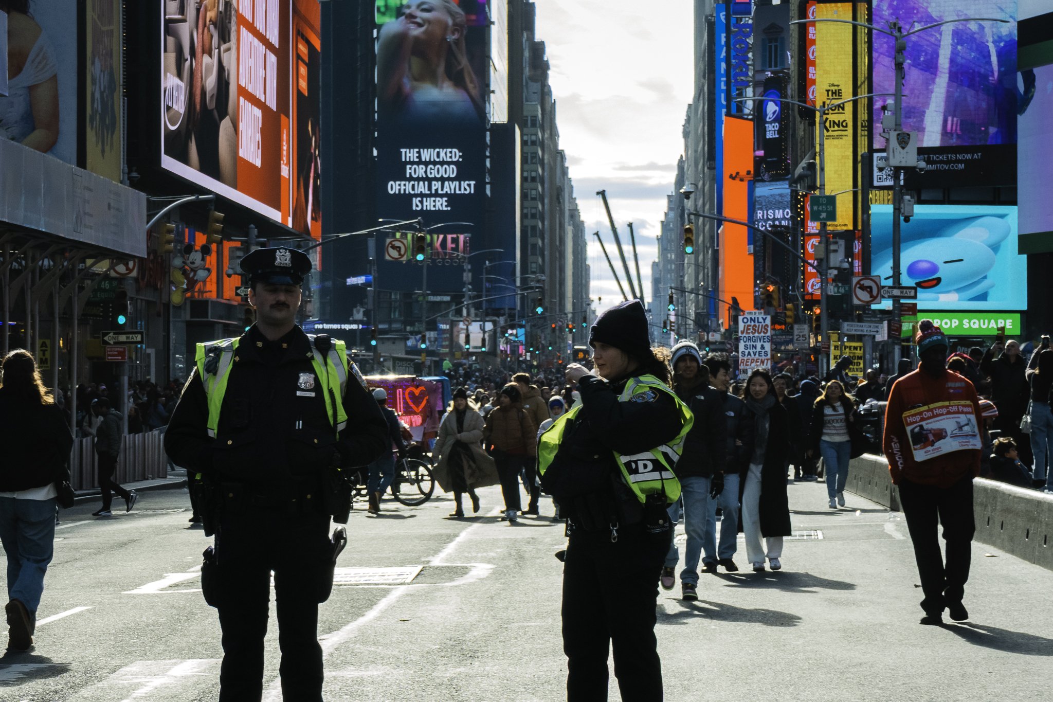 Crowded city street with many pedestrians, police officers in foreground, bright billboards and advertisements on buildings, some with neon lights, and a partly cloudy sky in the background.