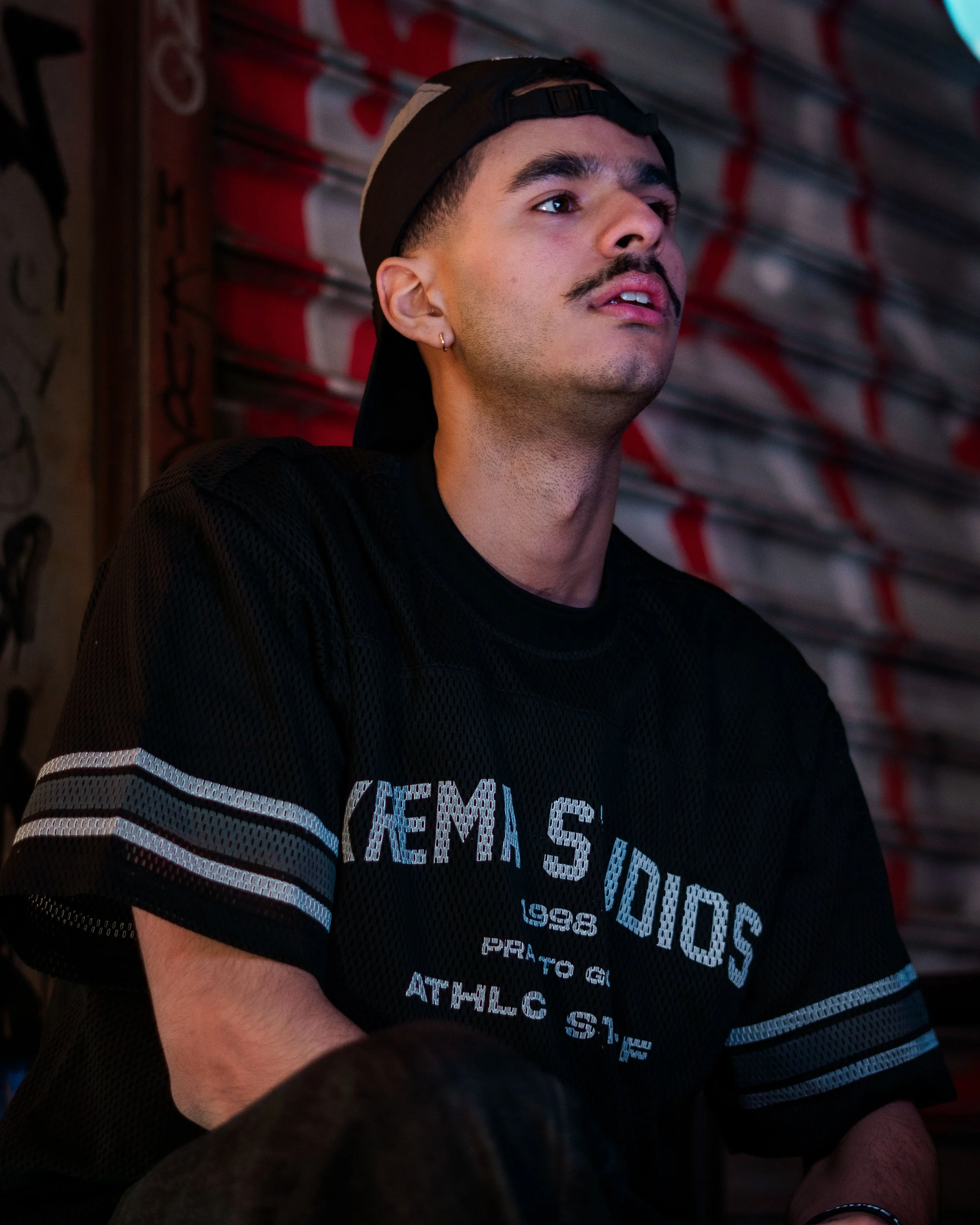 A young man with a mustache wearing a black cap backward and a black athletic shirt, looking upwards with a focused expression, sitting against a graffiti-covered wall.