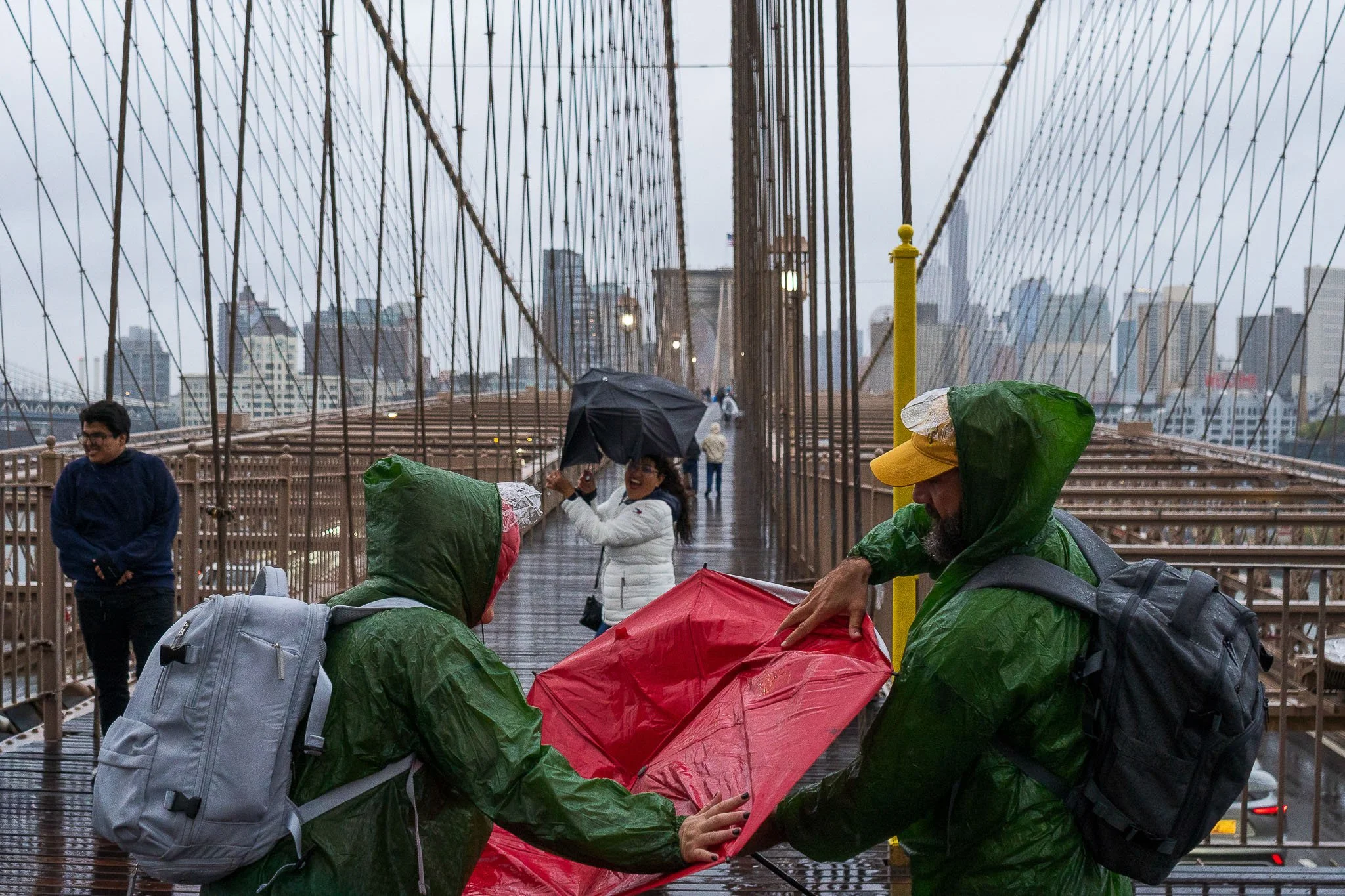 People on the Brooklyn Bridge in rainy weather, some wearing raincoats, holding umbrellas, and looking at a map.