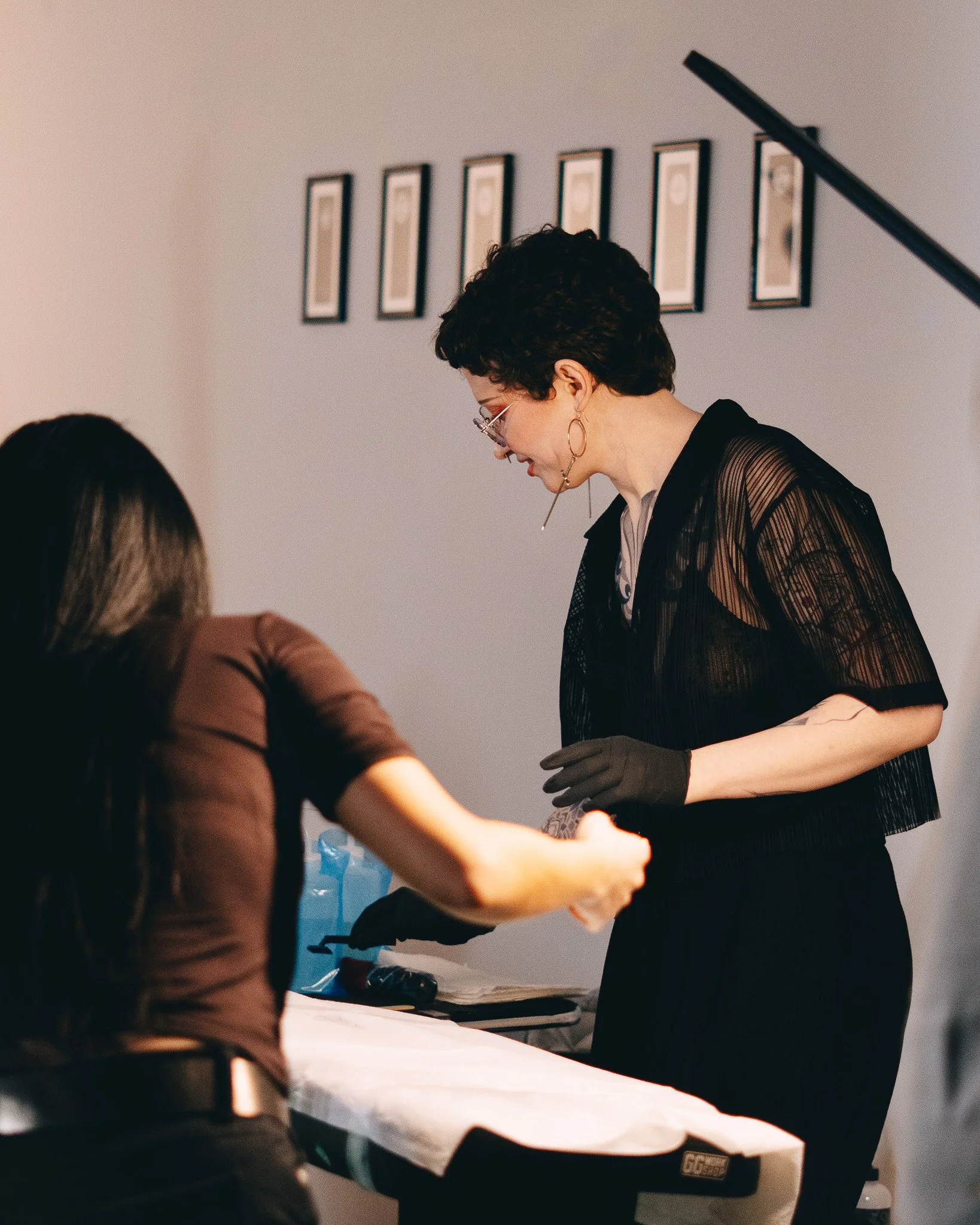 A woman with short dark hair, wearing glasses, earrings, black gloves, and a black top with sheer sleeves, is standing and interacting with another person who is seated at a white table. In the background, there are six framed pictures on the wall, a