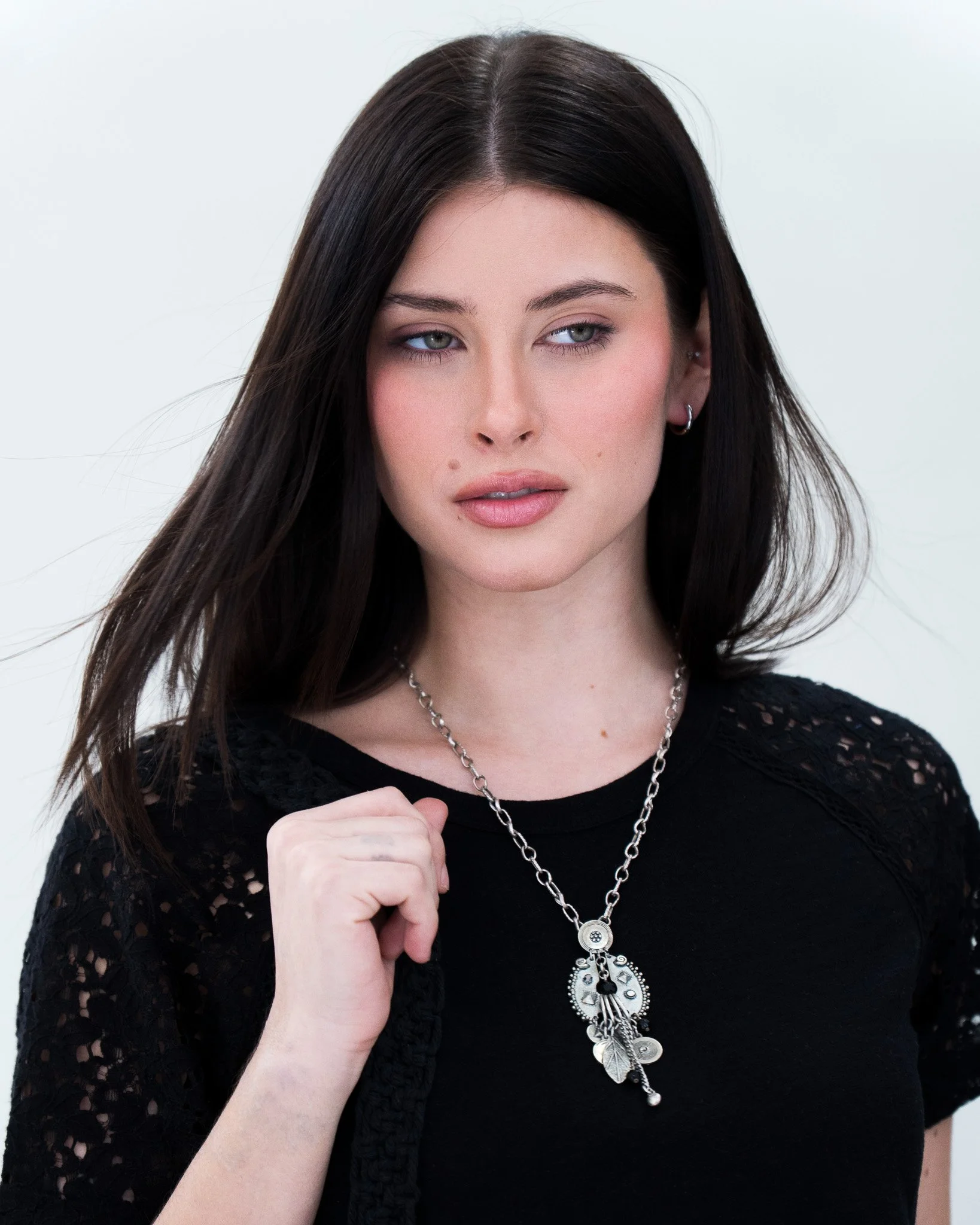 A woman with long dark hair wearing a black top with lace sleeves, a silver necklace with charms, and small hoop earrings. She is holding her fist near her chest in a confident pose.