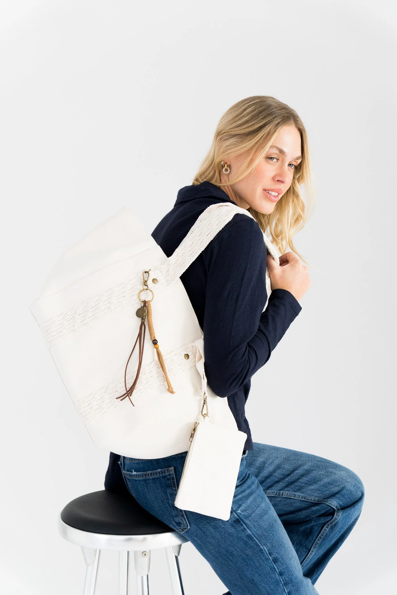 A woman sitting on a black stool with a white top, wearing a navy hoodie and blue jeans, carrying a large white tote bag over her shoulder, posing against a plain white background.