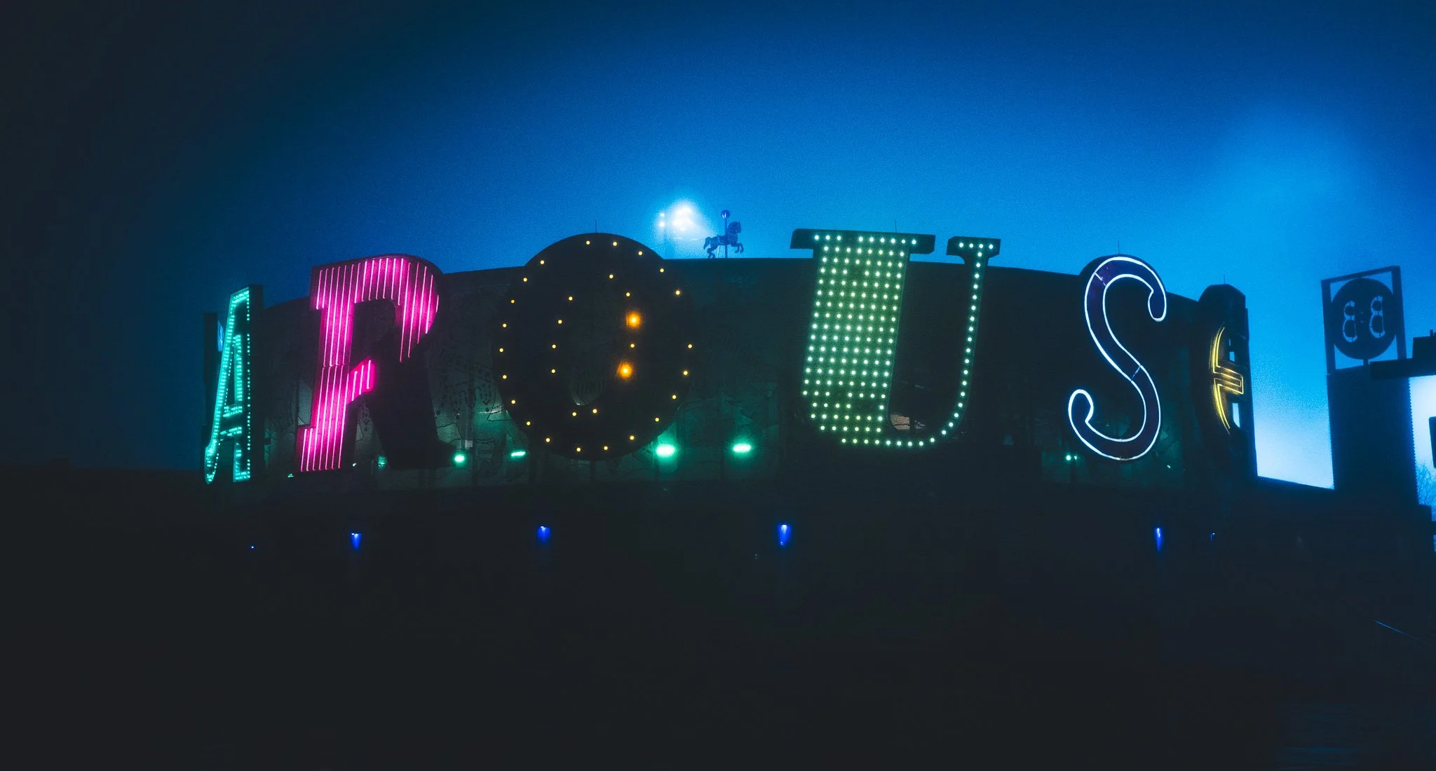 Nighttime view of a brightly lit amusement park ride with colorful neon lights spelling out the word 'FOLIES' on the building's facade, and a small silhouette of a knight on horseback atop a central part of the structure, against a dark sky.