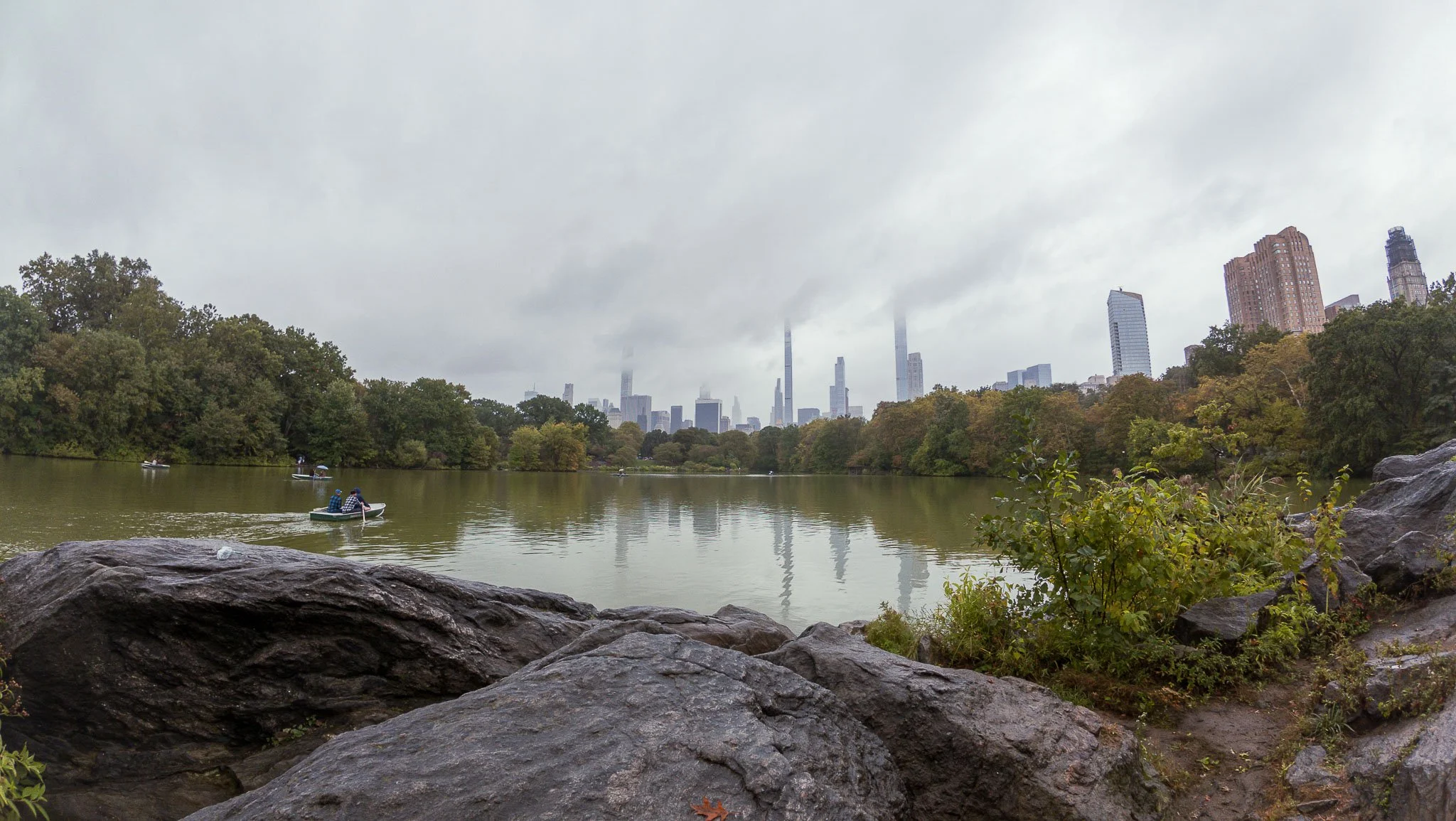 A view of a city skyline with skyscrapers shrouded in clouds, taken from a park with a lake and boats, and large rocks and greenery in the foreground on a cloudy day.