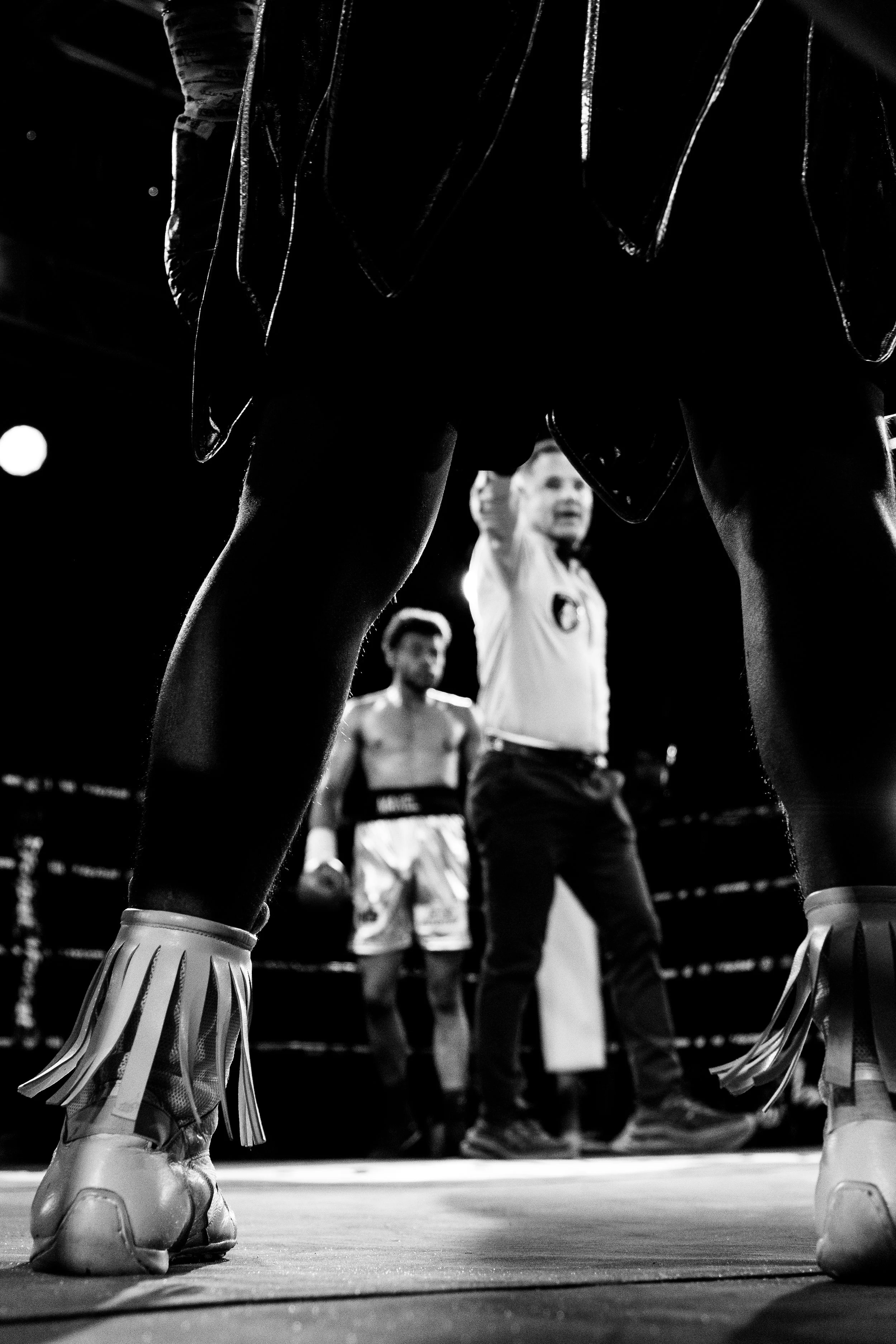 Boxing match seen from the perspective of the fighters' legs, showing the boxing ring, fighters' shorts, and shoes, with spectators and fighter in focus in the background.