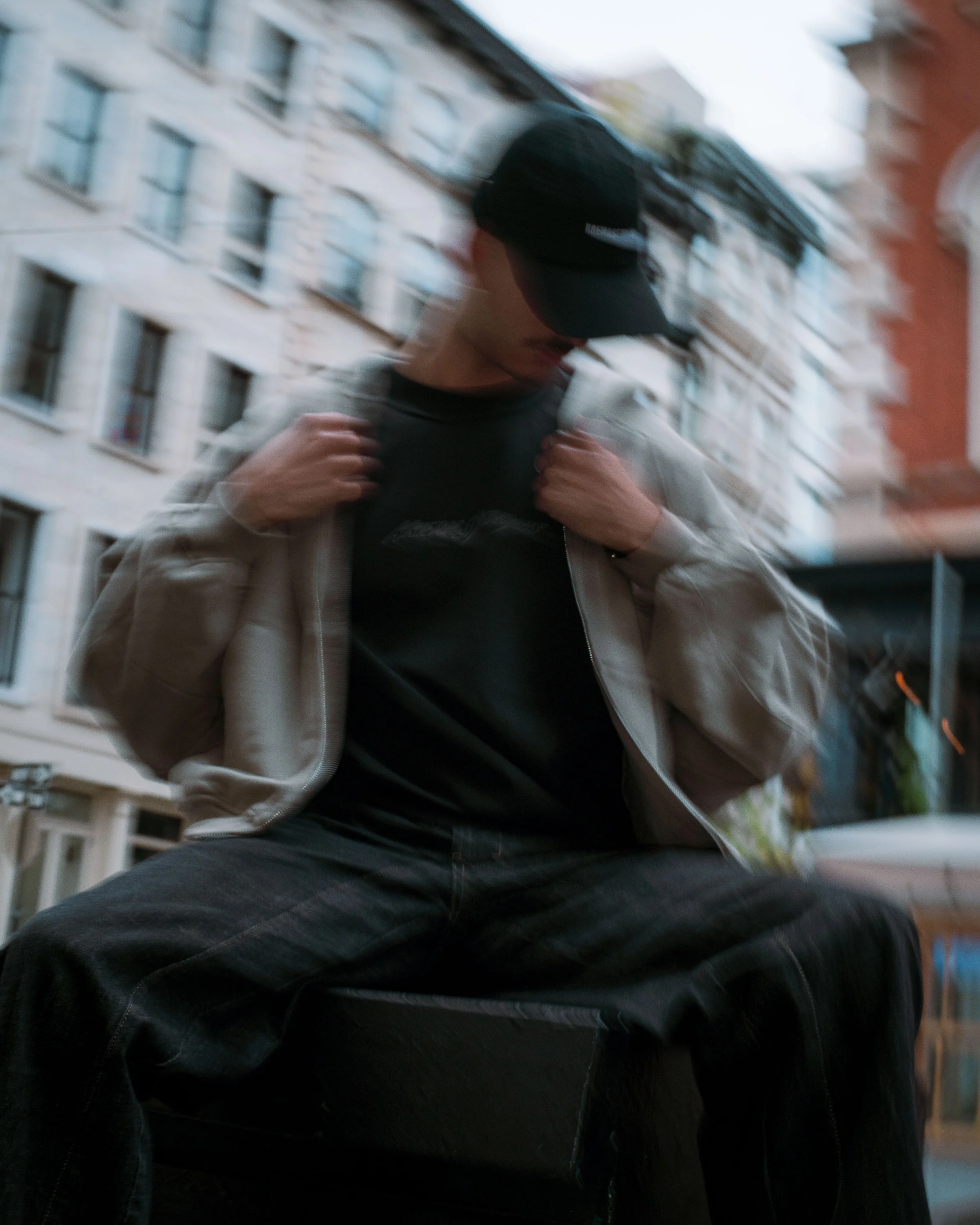 A person sitting outdoors in an urban area, wearing a black cap, dark clothing, and a light-colored jacket, with blurred city buildings in the background.