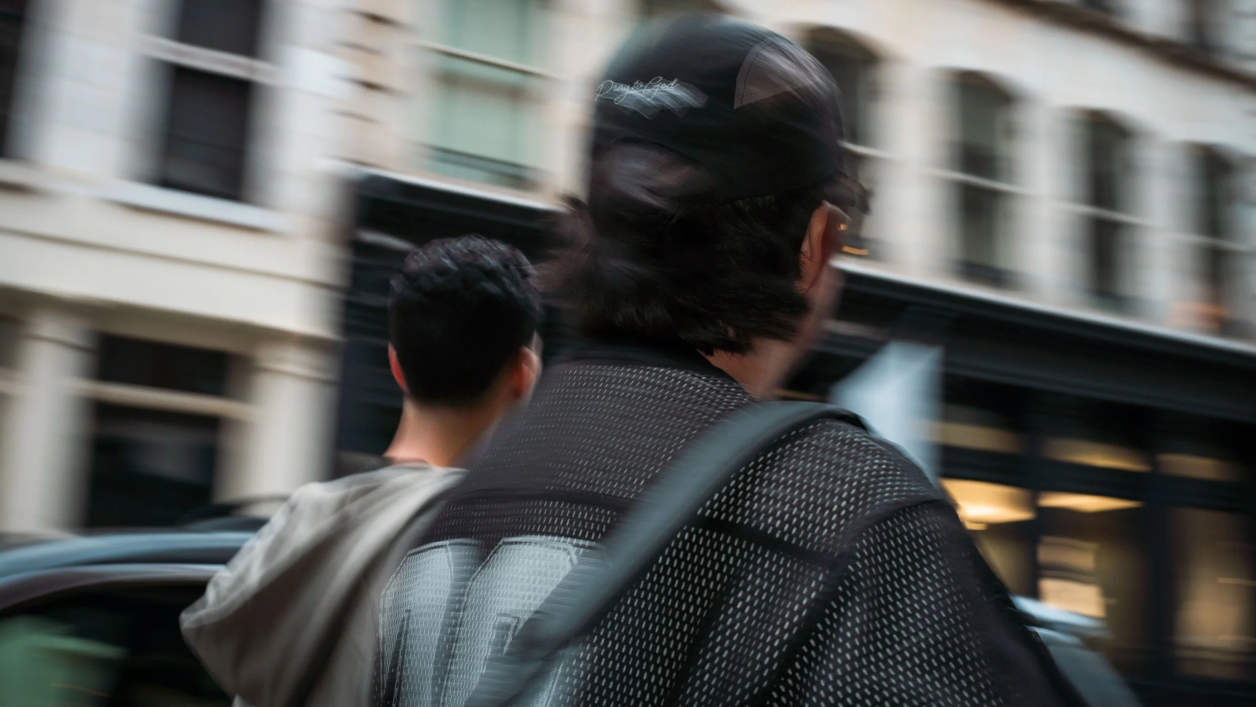 Two individuals walking outdoors in an urban setting with buildings in the background, one with a black cap and backpack, and the other with short dark hair, blurred motion.