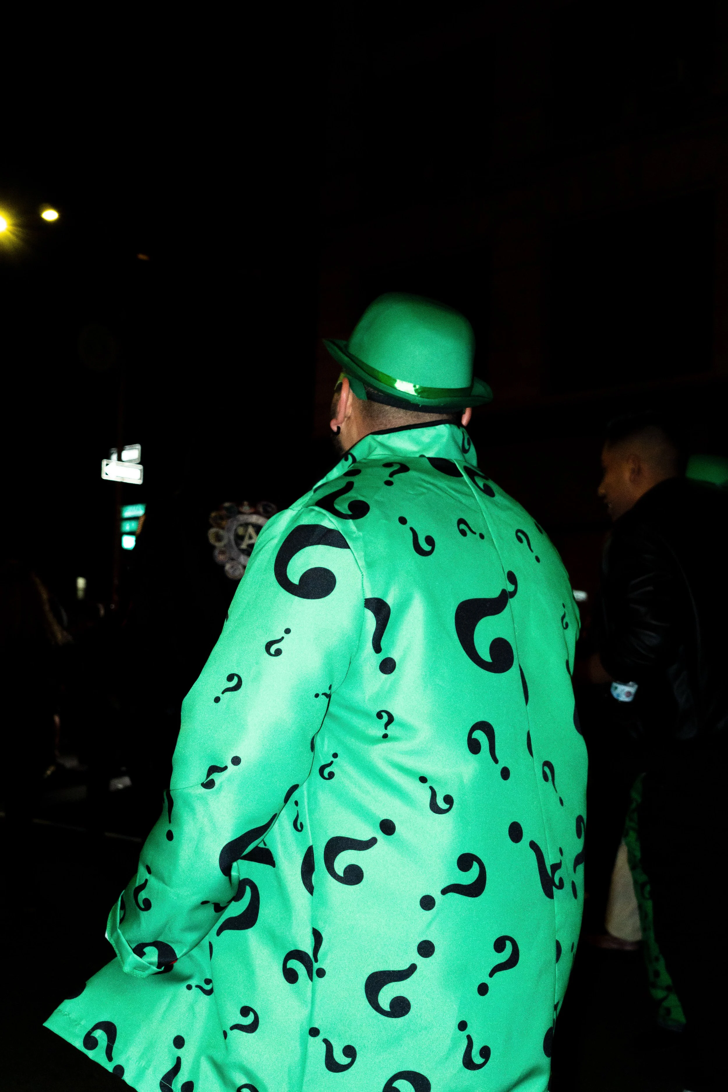 Man wearing a green hat and matching blazer with black question marks, illuminated in a dark setting.