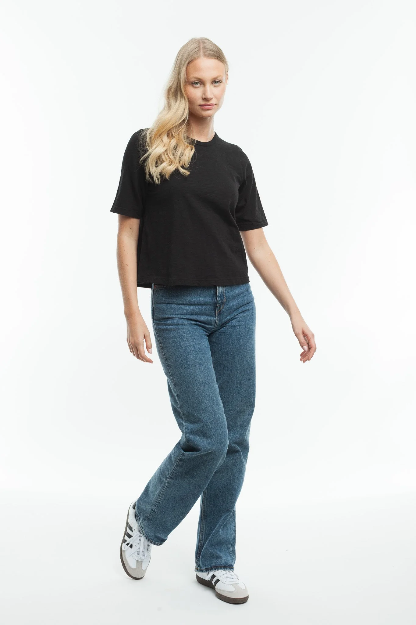 A young woman with long blonde wavy hair wearing a black t-shirt, blue jeans, and sneakers, standing against a plain white background.