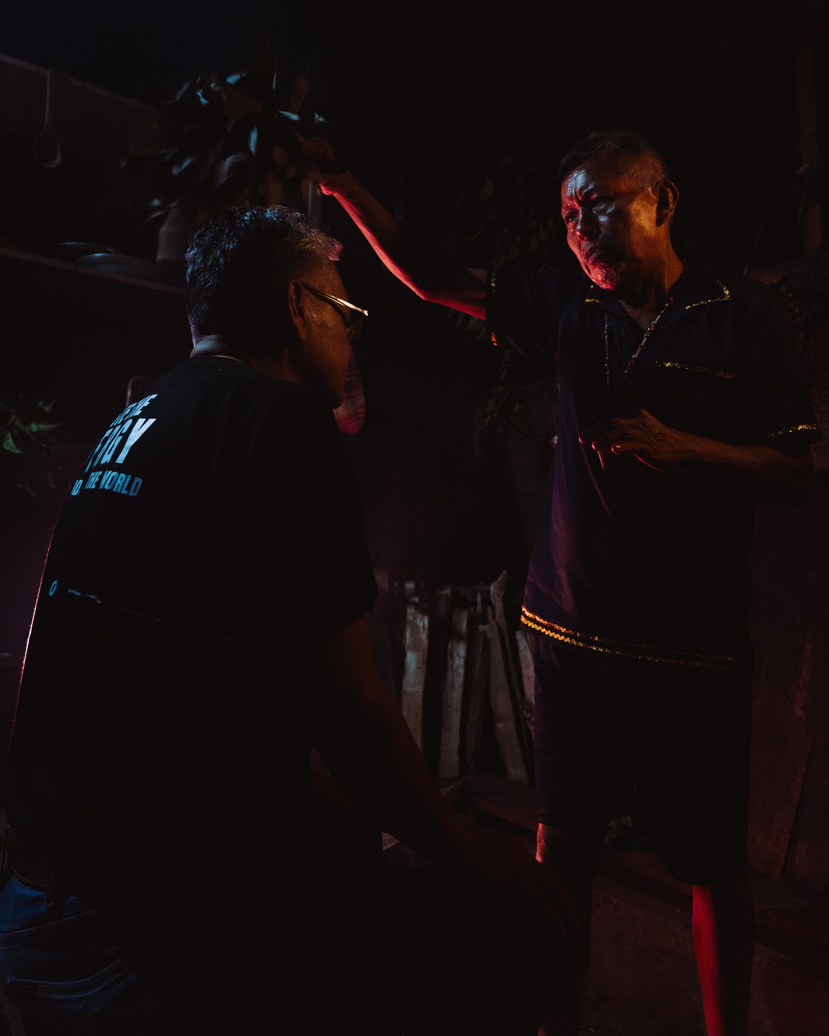 Two men having a conversation outdoors at night. One man is wearing glasses and a dark shirt, the other has short hair and a necklace, and is leaning on a red cane or stick. The background is dark, with partially visible trees.