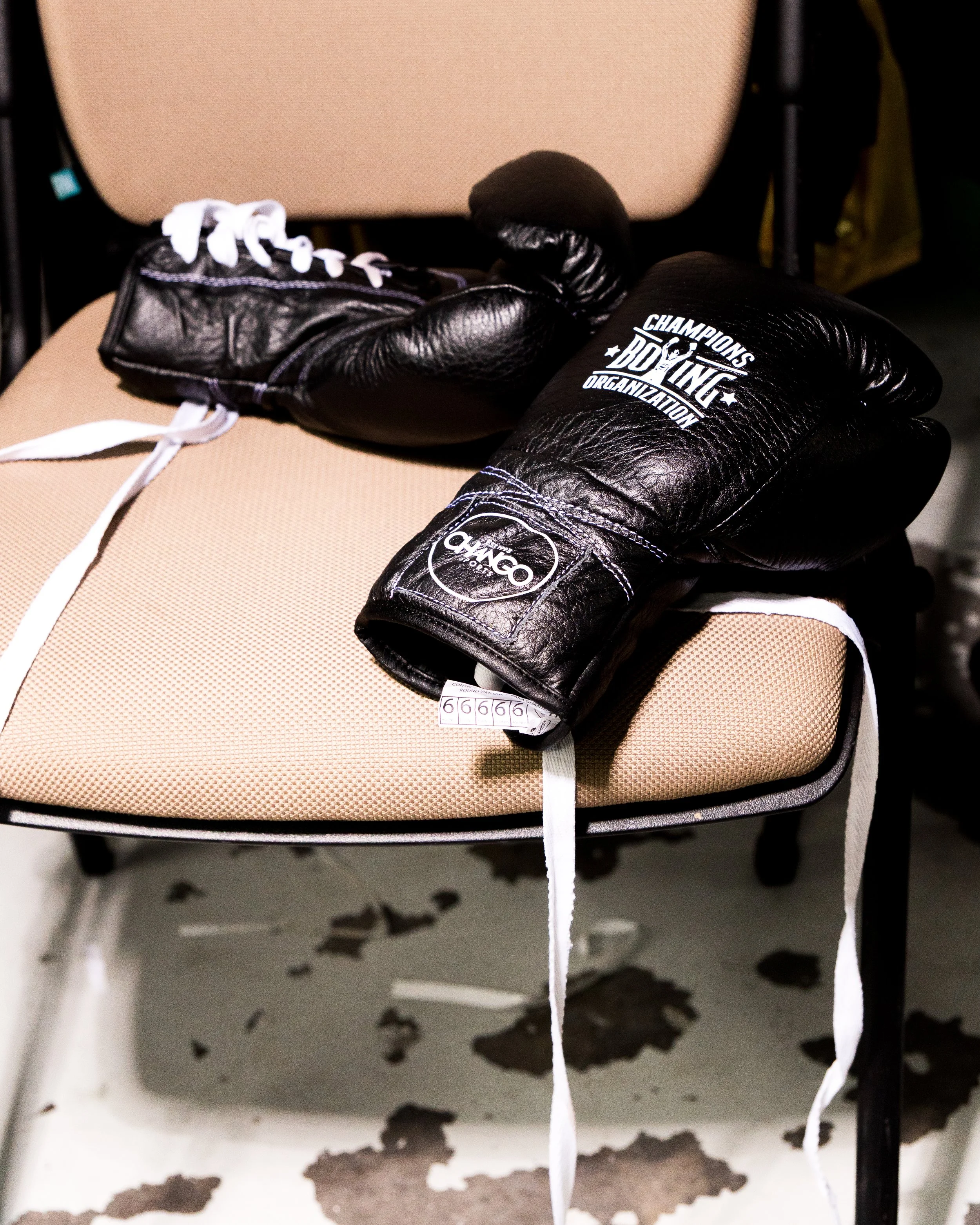 A beige-colored chair with a pair of black boxing gloves resting on it, with white laces and a logo that reads 'Champions Boxing Organization.' The background shows a wall with chipped paint and black marks.