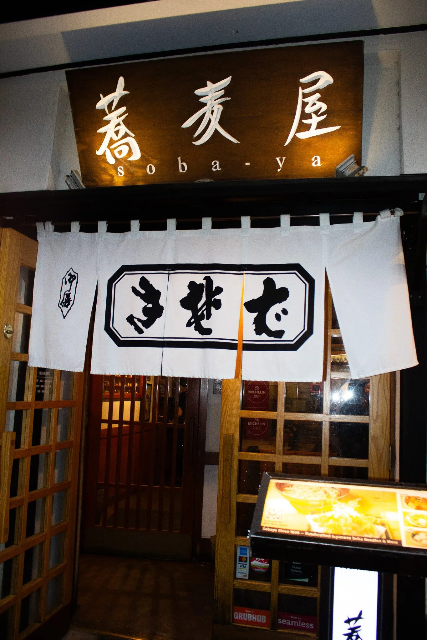 Restaurant entrance with a wooden sign reading 'soba-ya' and traditional white noren curtains with Japanese characters, featuring a menu display stand outside.