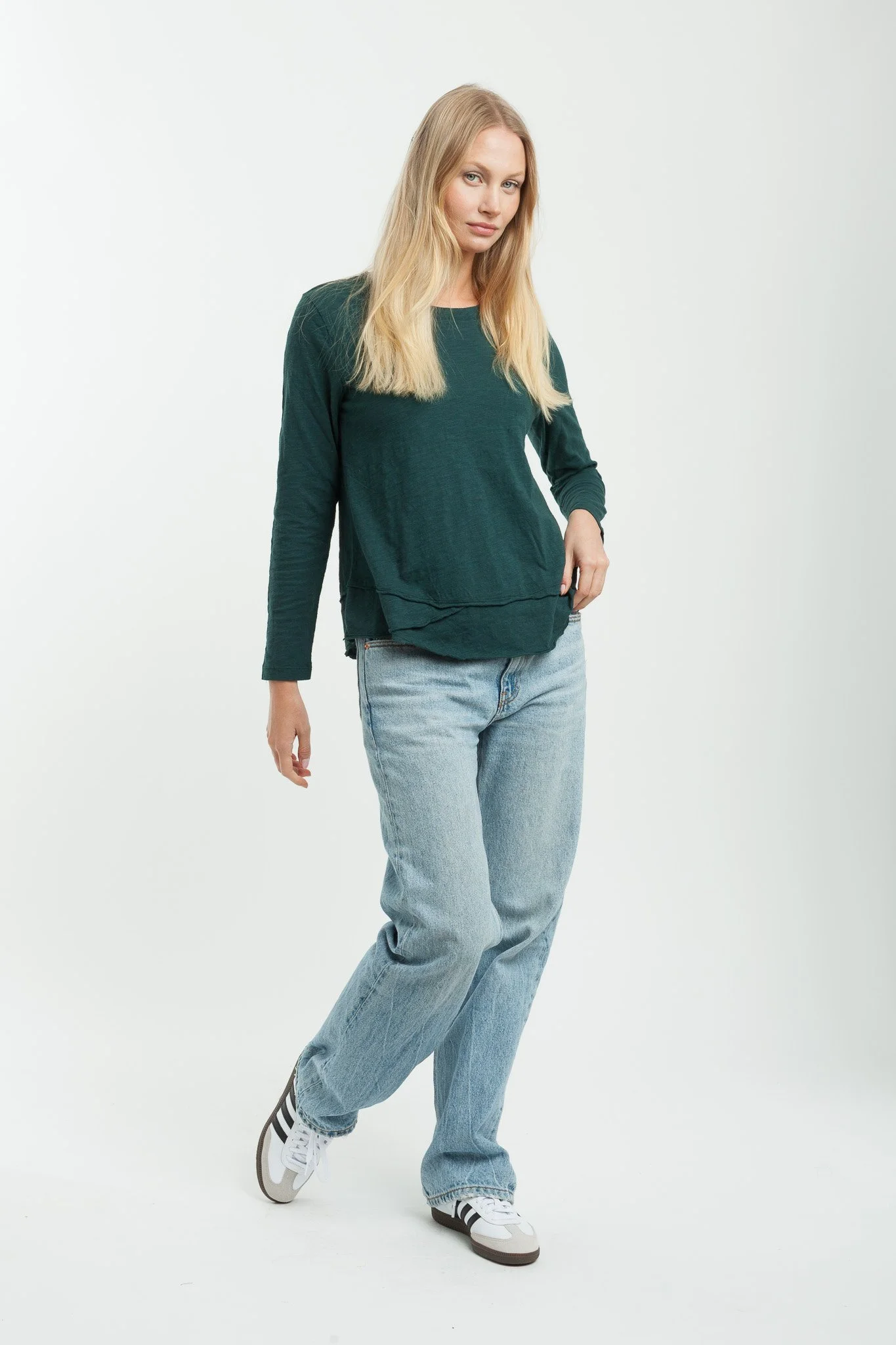 Woman with long blonde hair wearing a dark green long-sleeve shirt, light blue jeans, and white sneakers with black stripes, standing against a plain white background.