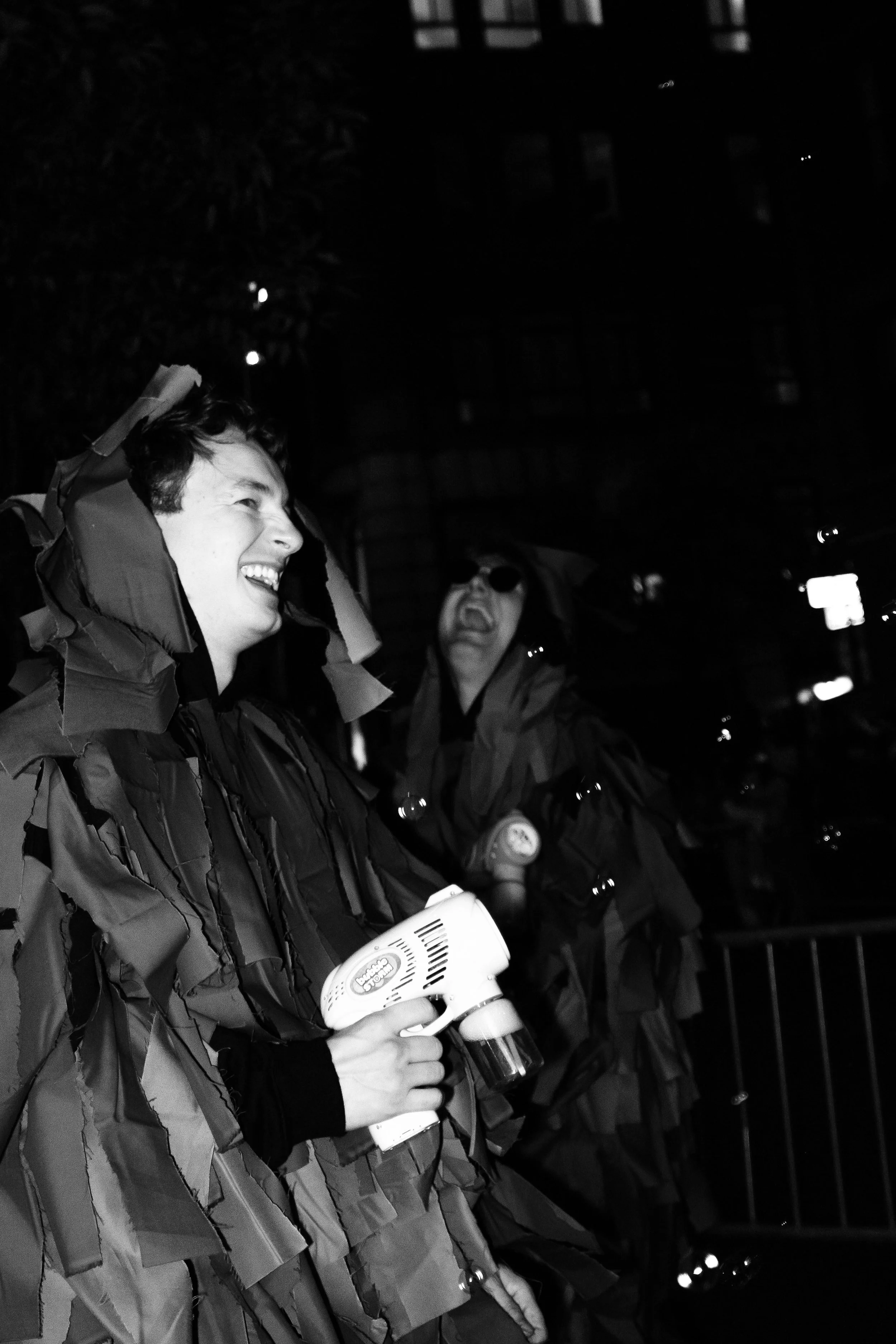 Two people laughing outdoors at night in raincoats, one holding a hairdryer, with dark background and faint city lights.