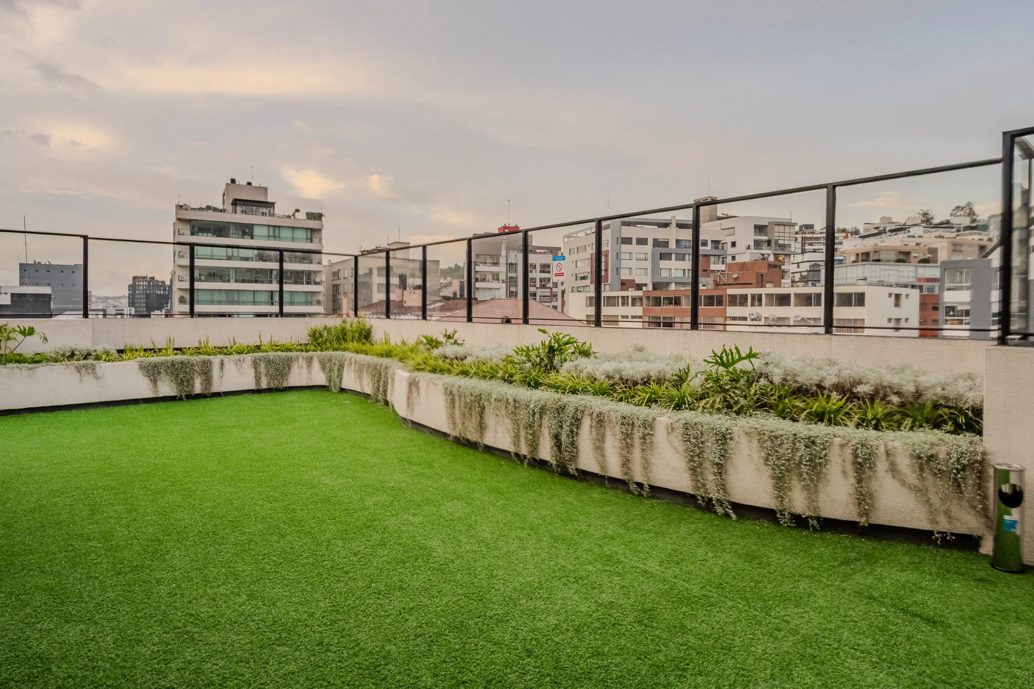 A rooftop garden with artificial grass and raised flower beds, surrounded by a black metal railing, overlooking a cityscape of modern apartment buildings.