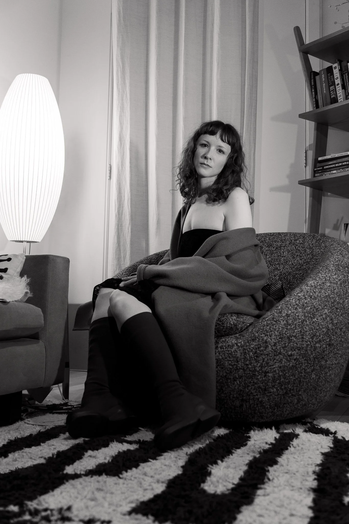 A woman with curly hair sitting on a sofa in a living room, with a patterned rug, a bookshelf with books, and a floor lamp nearby.