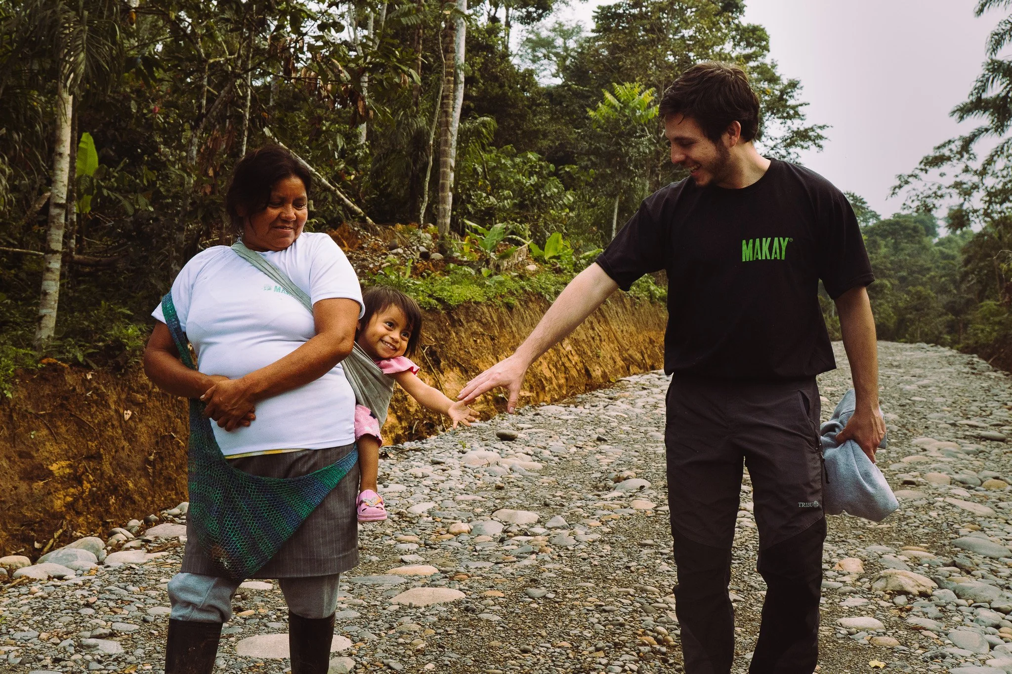 Two people and a child walking along a rocky riverbank surrounded by lush green trees. One person is helping the child cross the river, and they are all smiling.