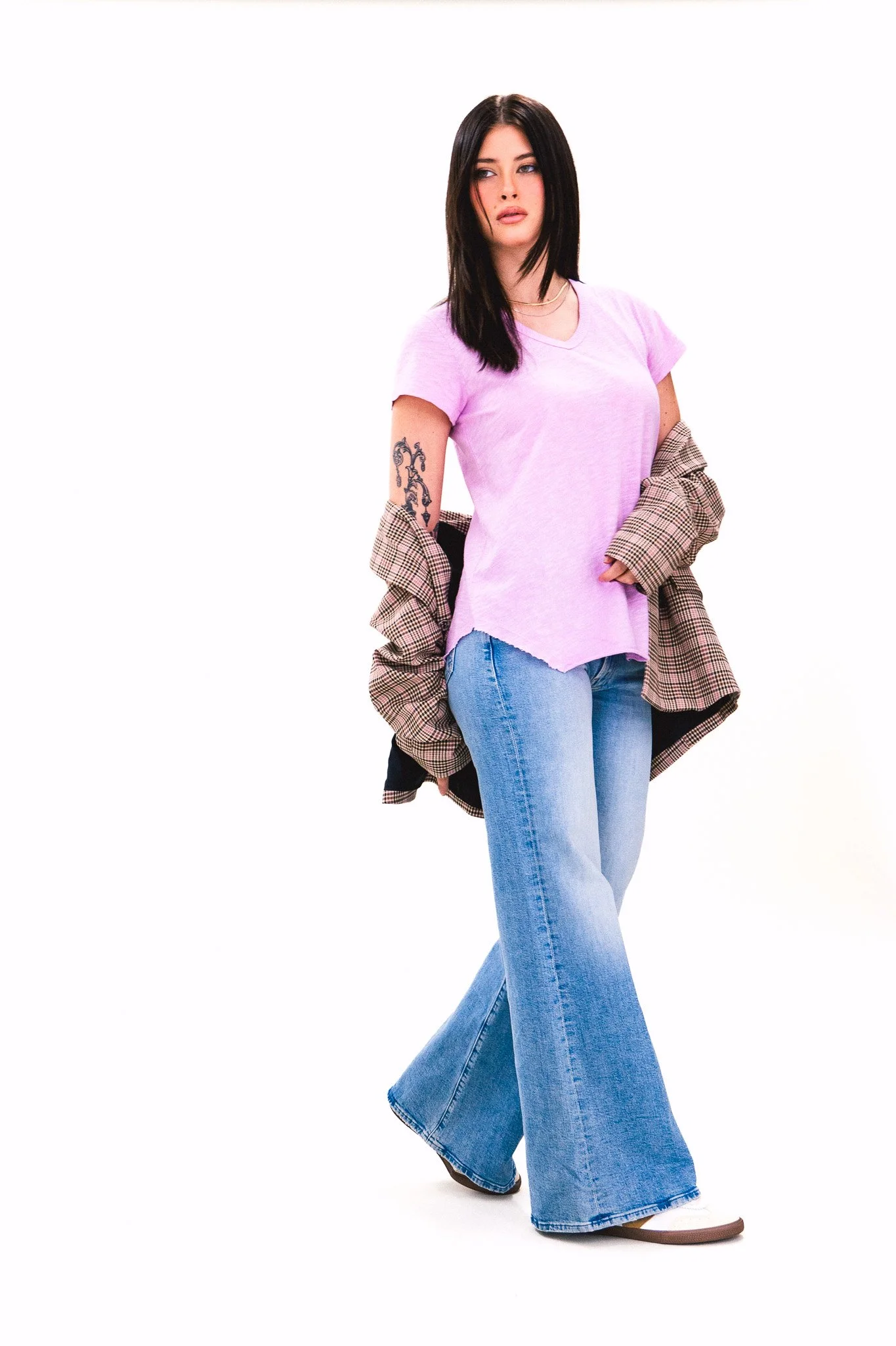A young woman with black hair wearing a light purple T-shirt, a plaid jacket draped over her shoulders, and blue wide-leg jeans with brown shoes. She has a tattoo on her left arm and is standing against a white background.