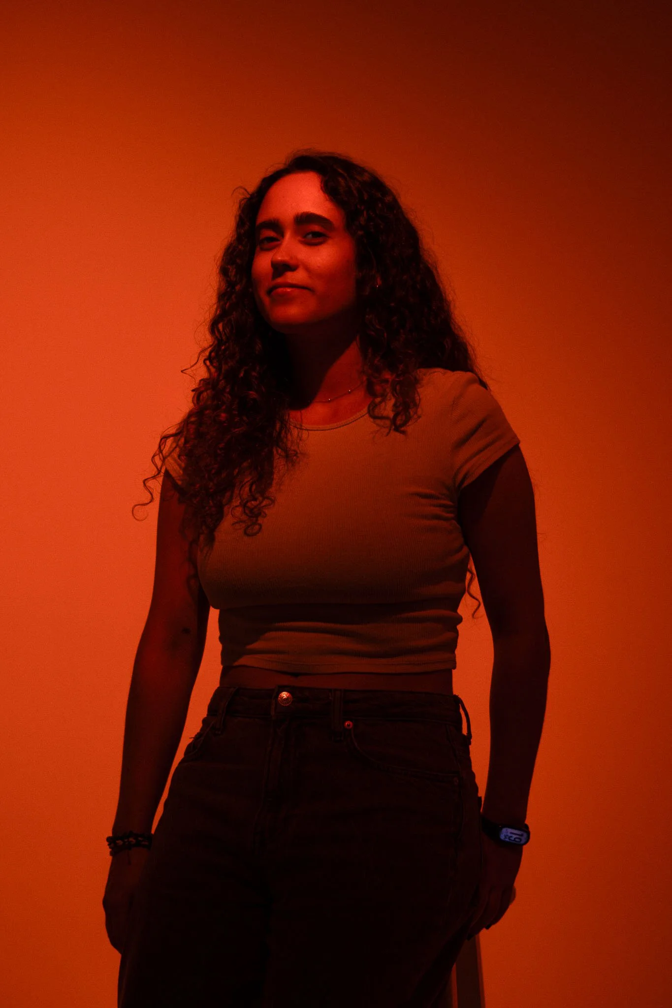 A woman with curly hair standing against an orange background, wearing a yellow t-shirt, dark pants, a necklace, and a watch.