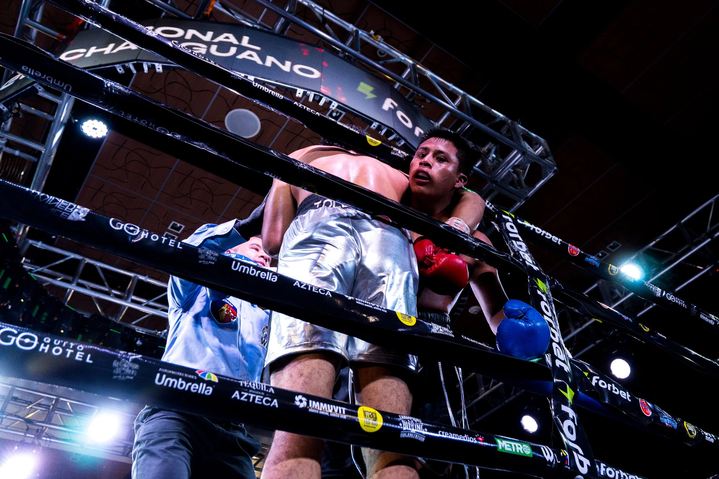 Two boxers in a boxing ring, with one leaning over the other's back, and a referee holding the leg of the boxer with red gloves. The scene takes place indoors with bright lighting and a large struggle between the fighters.