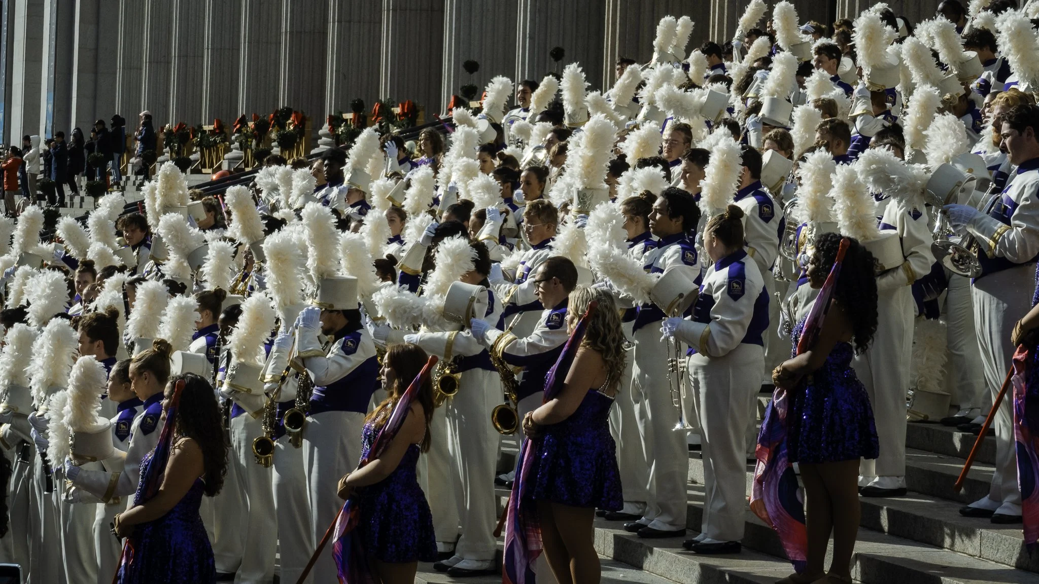 A large group of marching band members dressed in white uniforms with blue accents, wearing tall white feathered hats, holding musical instruments, and assembled outdoors on steps during a parade or event.