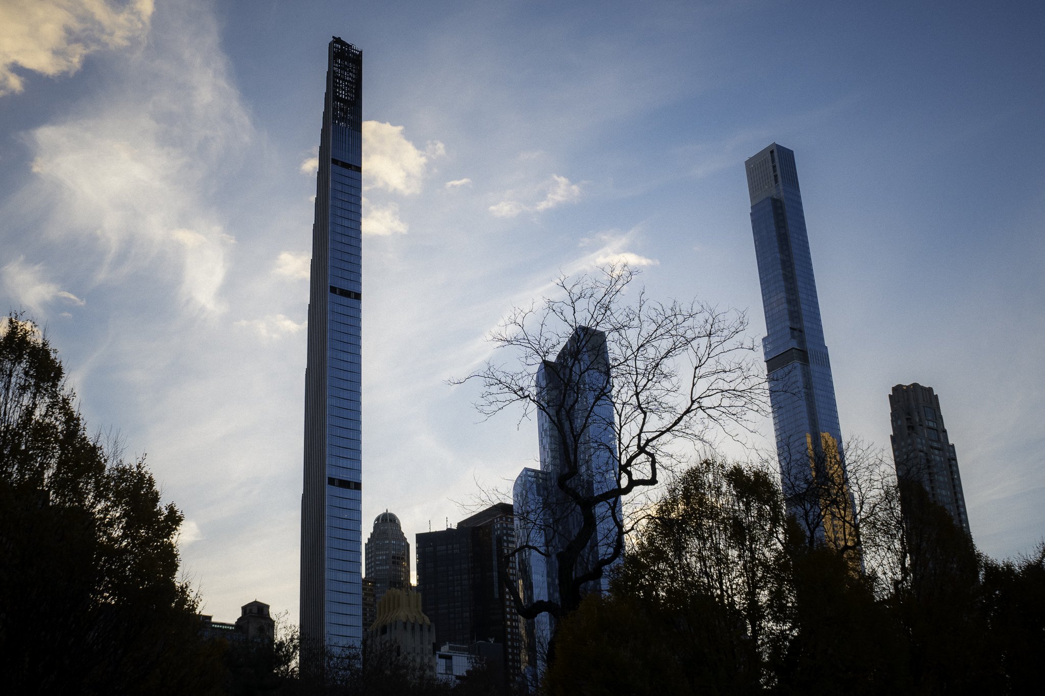 View of several tall modern skyscrapers in a city, with leafless trees in the foreground and a partly cloudy sky in the background.