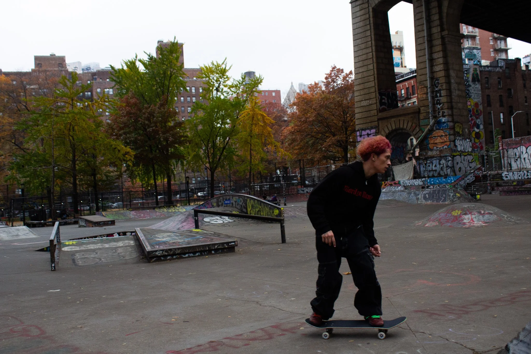 Person skateboarding at an outdoor skatepark during overcast weather with graffiti-covered ramps and trees with autumn foliage.