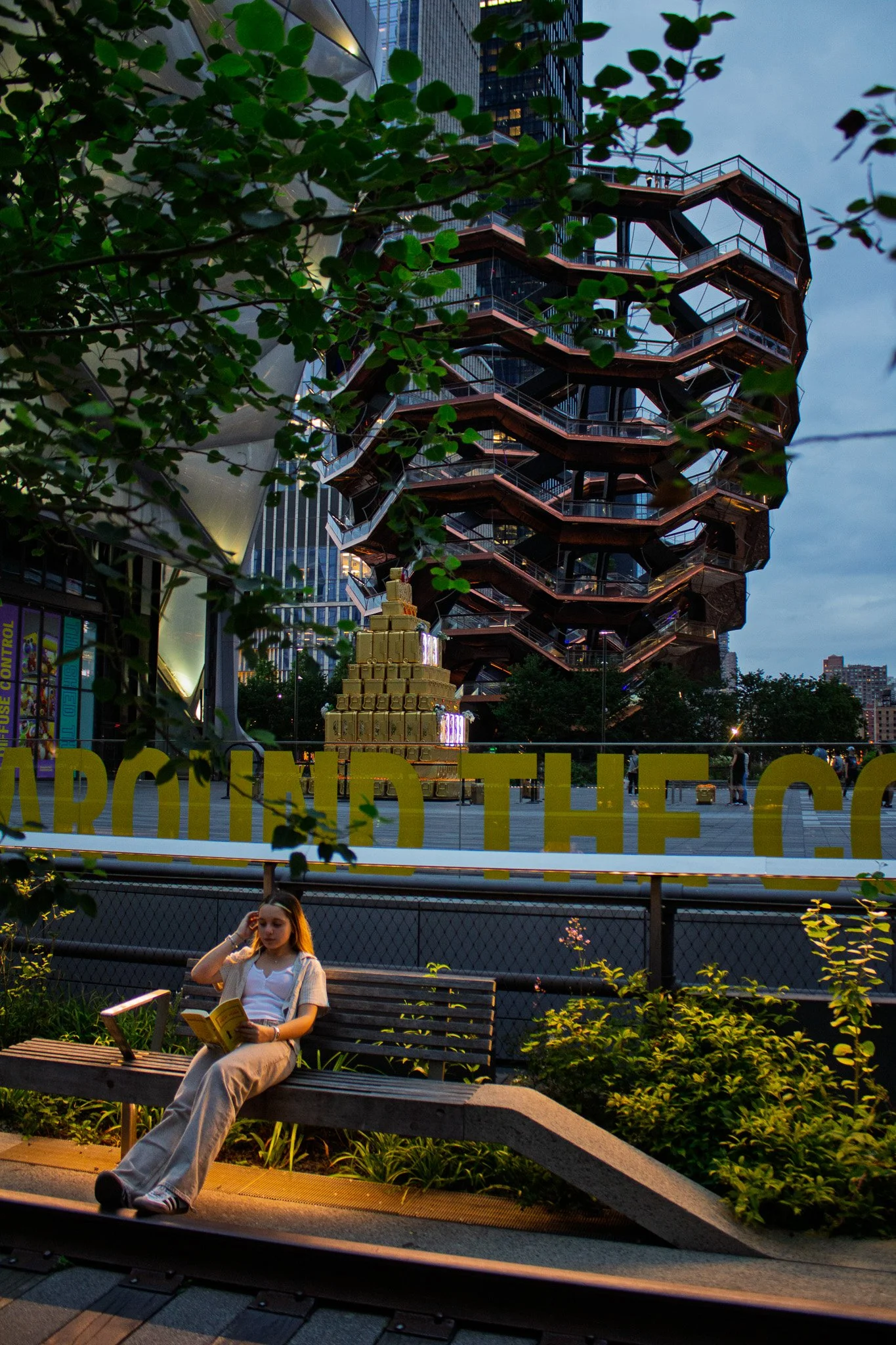 A young woman is sitting on a park bench, reading a book, in front of a large modern building with an outdoor staircase structure, surrounded by green plants and trees, during evening hours.