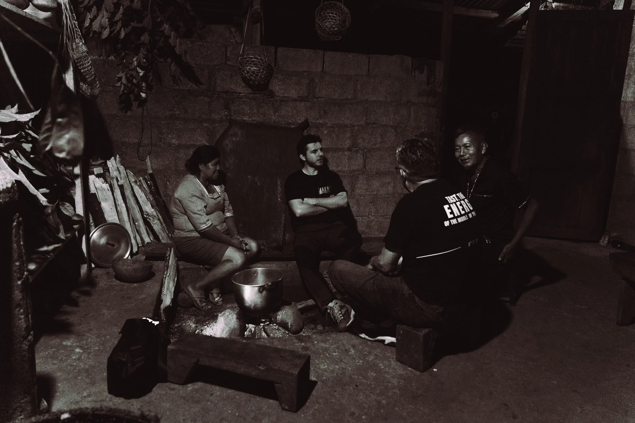 A group of five people sitting in a corner of a rustic room, engaged in conversation. The room has a brick wall with hanging baskets, a shelf with books and fabrics, and a fire pit on the floor.