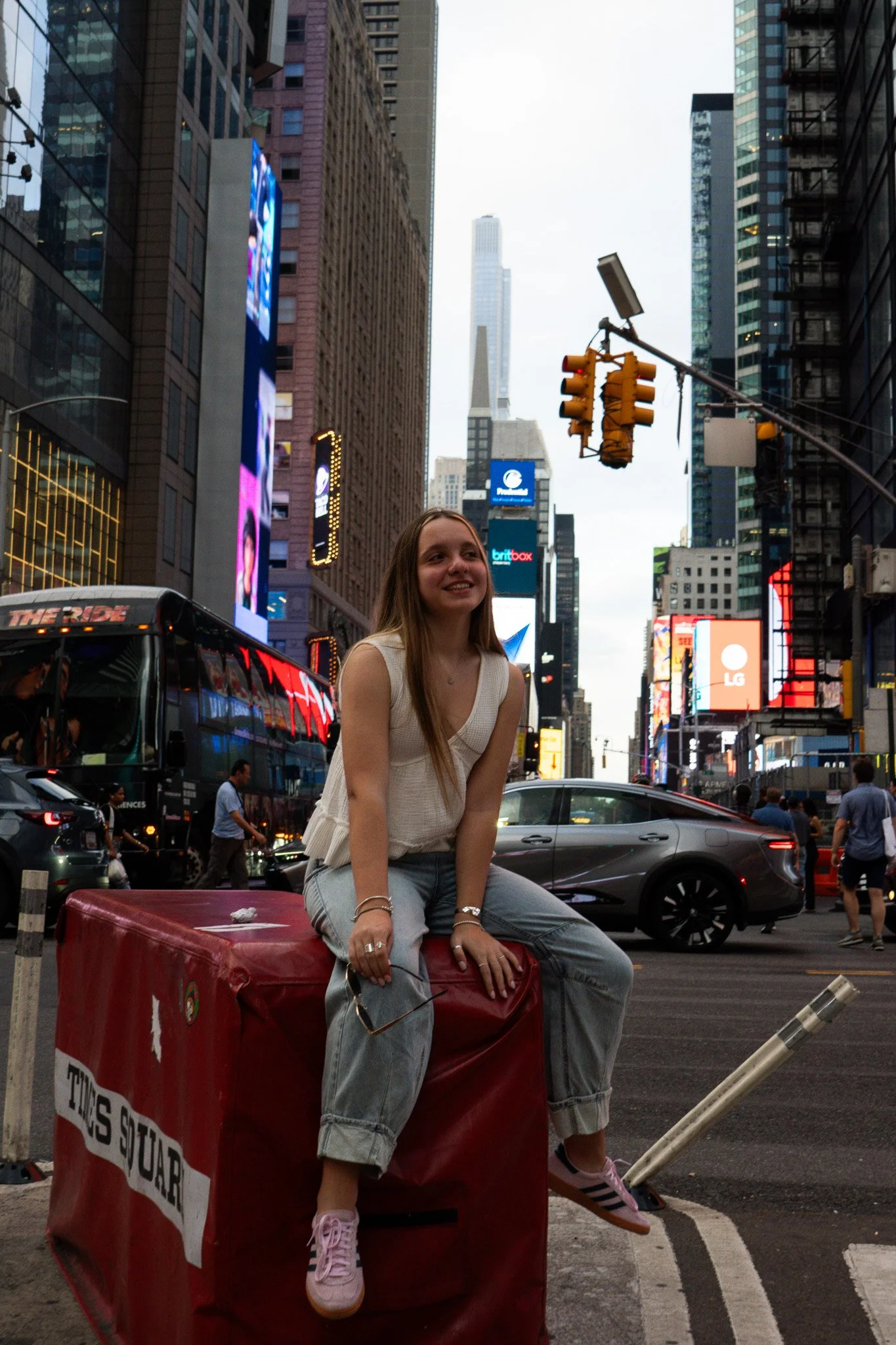 A young woman sitting on a red trash bin in Times Square, New York City, with tall buildings, electronic billboards, and traffic around her.