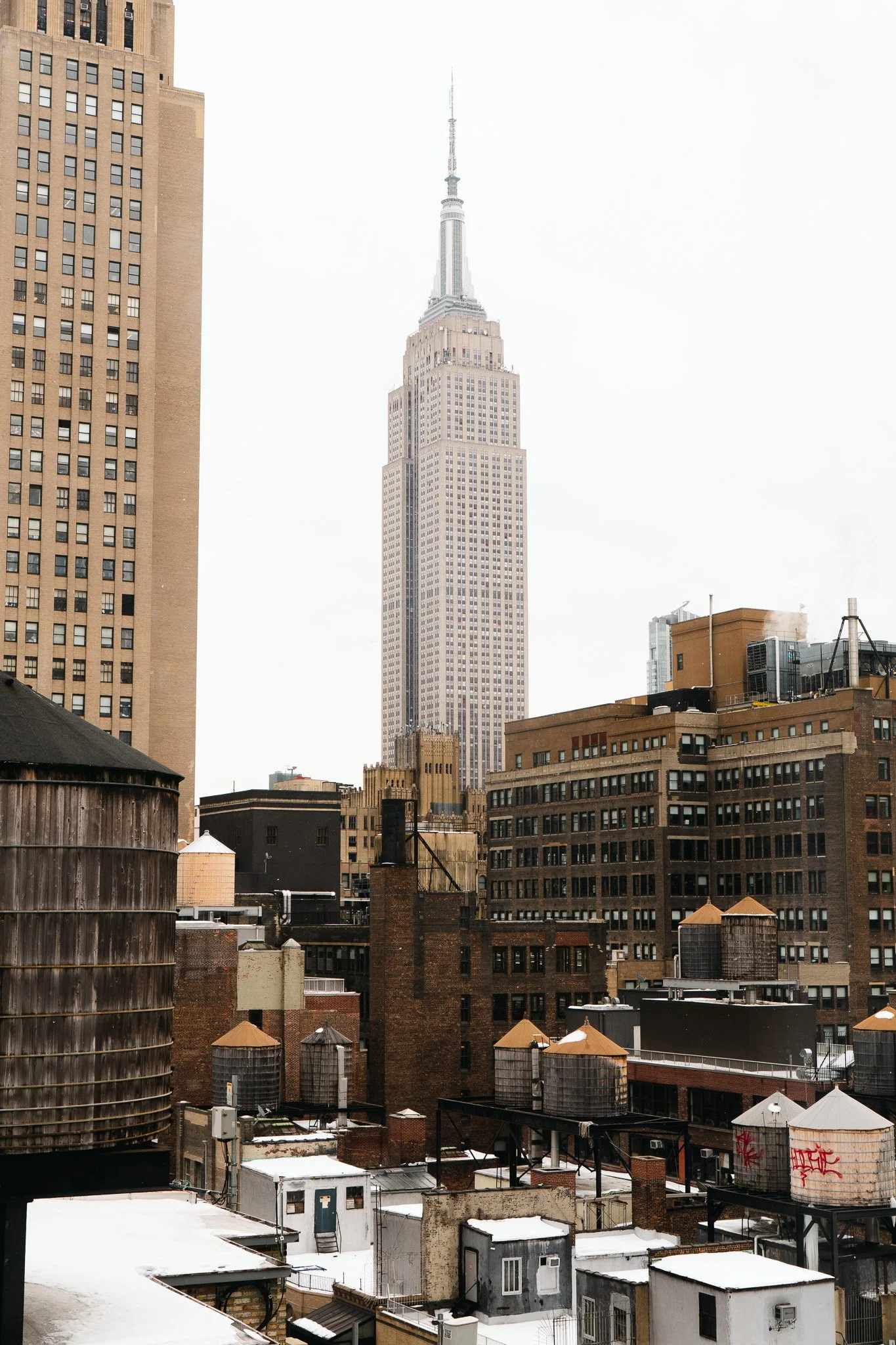 City skyline with various buildings and water towers on rooftops, including the Empire State Building in the background, under cloudy sky.